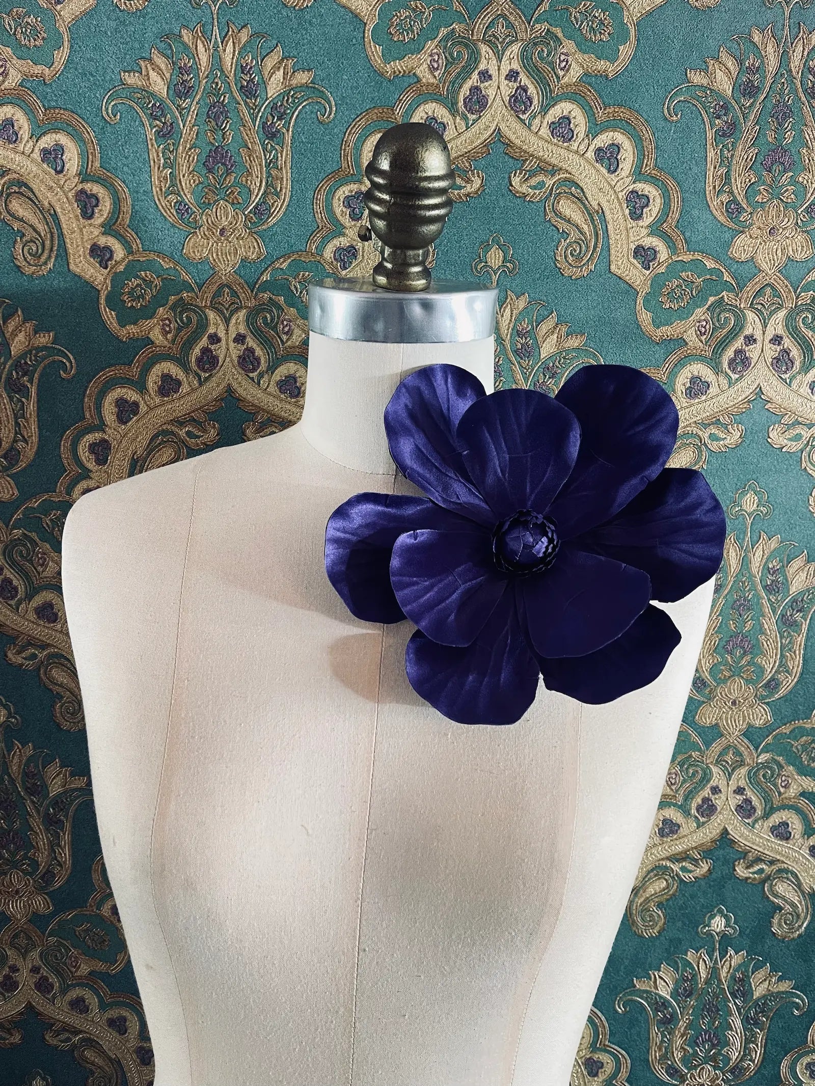 Broochella Magnolia handmade large flower brooch in purple. Styled on a mannequin. The background is green and gold damask wallpaper