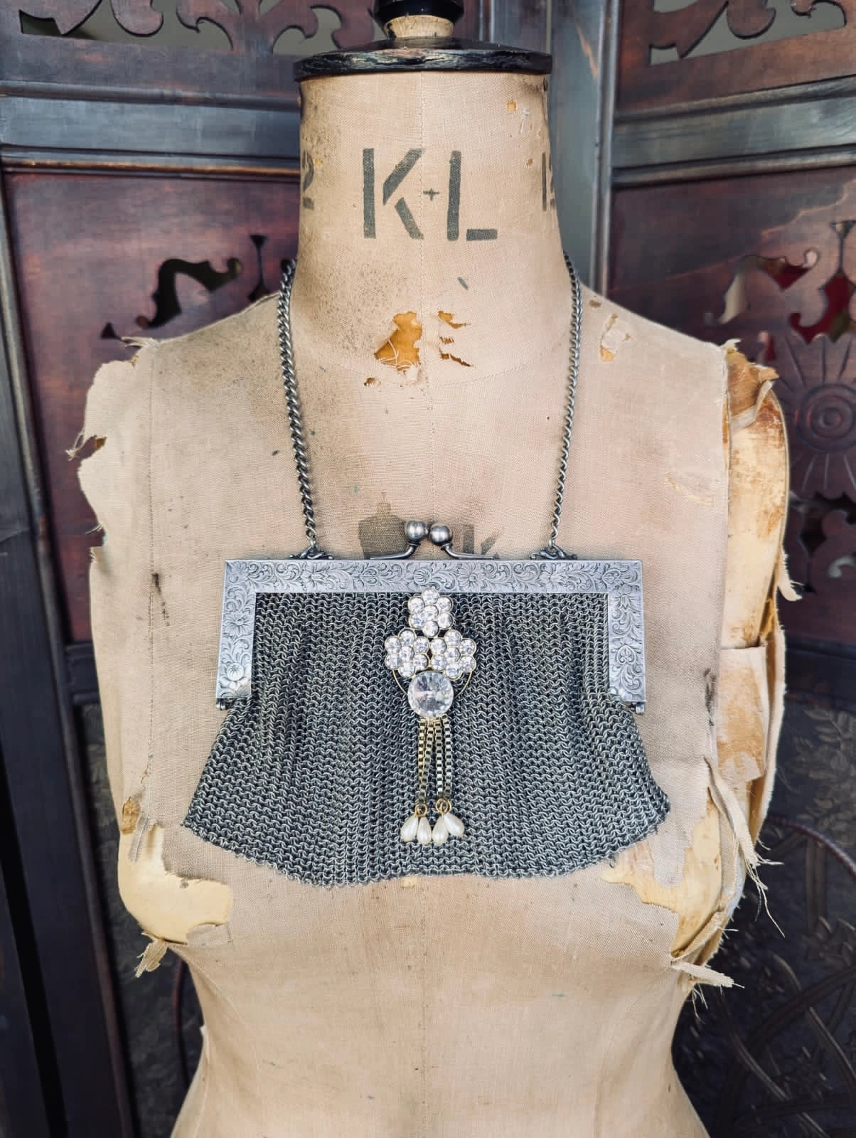 A vintage Broochella maison broche bijou argente silver chain mesh clutch purse with a textured silver-tone frame, brooch embellishment , and dangling pearl tassel. It is displayed on a vintage mannequin. The background is a decorative, brown, wooden panel. 