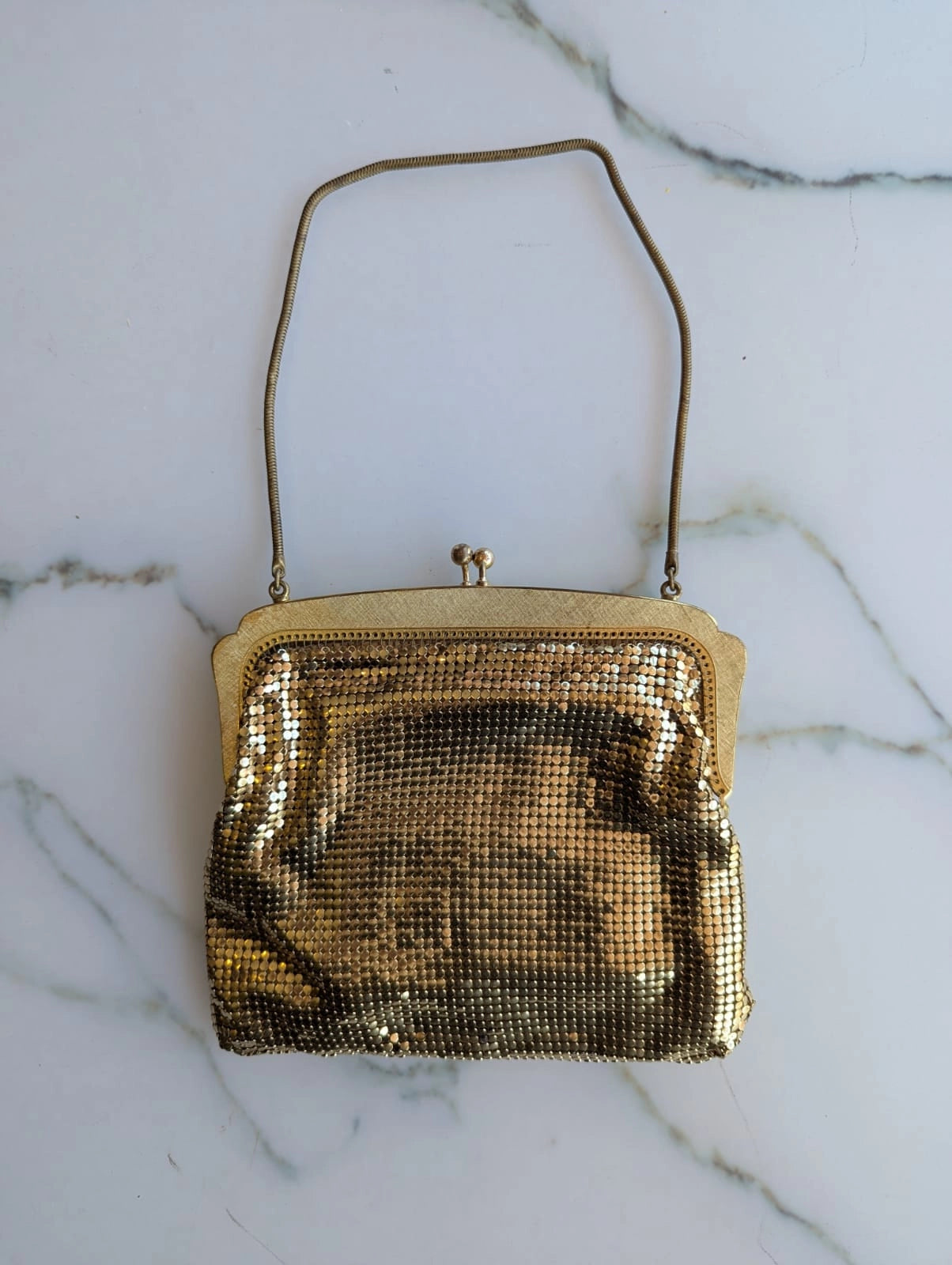 A Broochella etoile d'or embellished vintage gold-tone mesh Oroton clutch purse with a curved shoulder strap, featuring a central cameo-style oval portrait, floral embellishments, and a bird motif, displayed on a white marble surface.