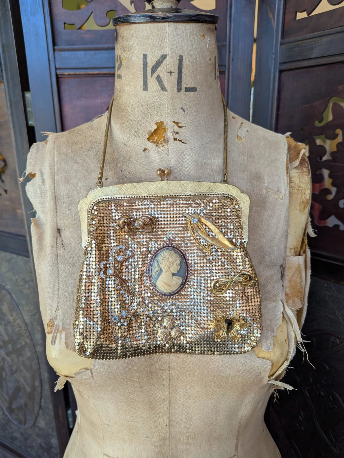 A Broochella etoile d'or embellished vintage gold-tone mesh Oroton clutch purse with a curved shoulder strap, featuring a central cameo-style oval portrait, floral embellishments, and a bird motif. It is displayed on a vintage mannequin. The background is a decorative brown wooden screen. 