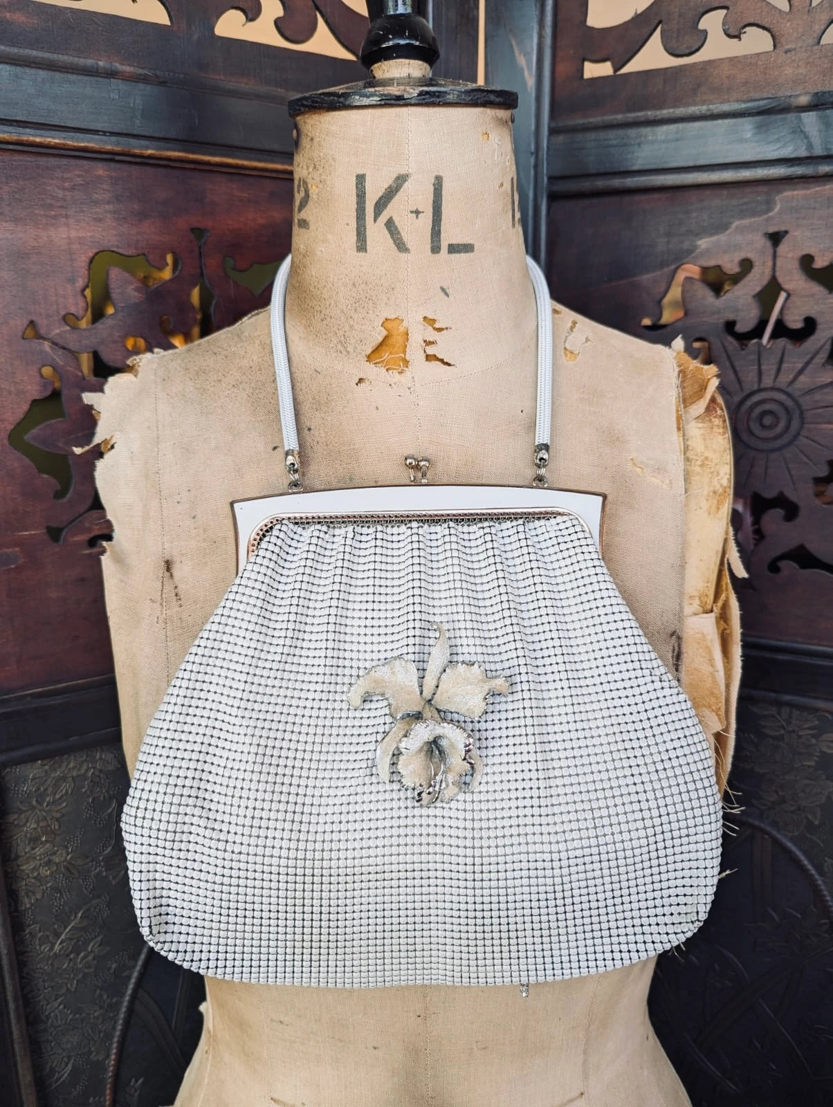 A Broochella Maison Broche l'orchidee vintage white glomesh clutch purse with a white floral brooch and white shoulder straps, displayed on a tan fabric mannequin torso against a dark carved wooden background.
