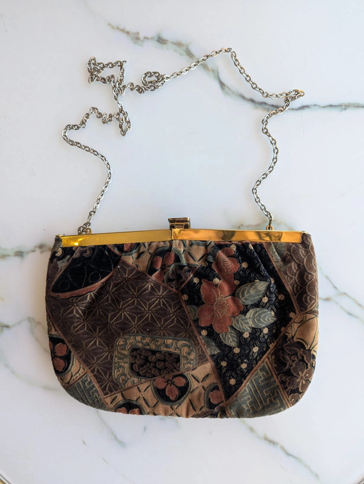 A Broochella Maison Broche la cameo du jardin vintage clutch handbag with a floral and geometric patchwork pattern in brown, black, and muted red, featuring a gold-tone metal frame and a silver-tone chain strap, laid flat on a white marble surface.