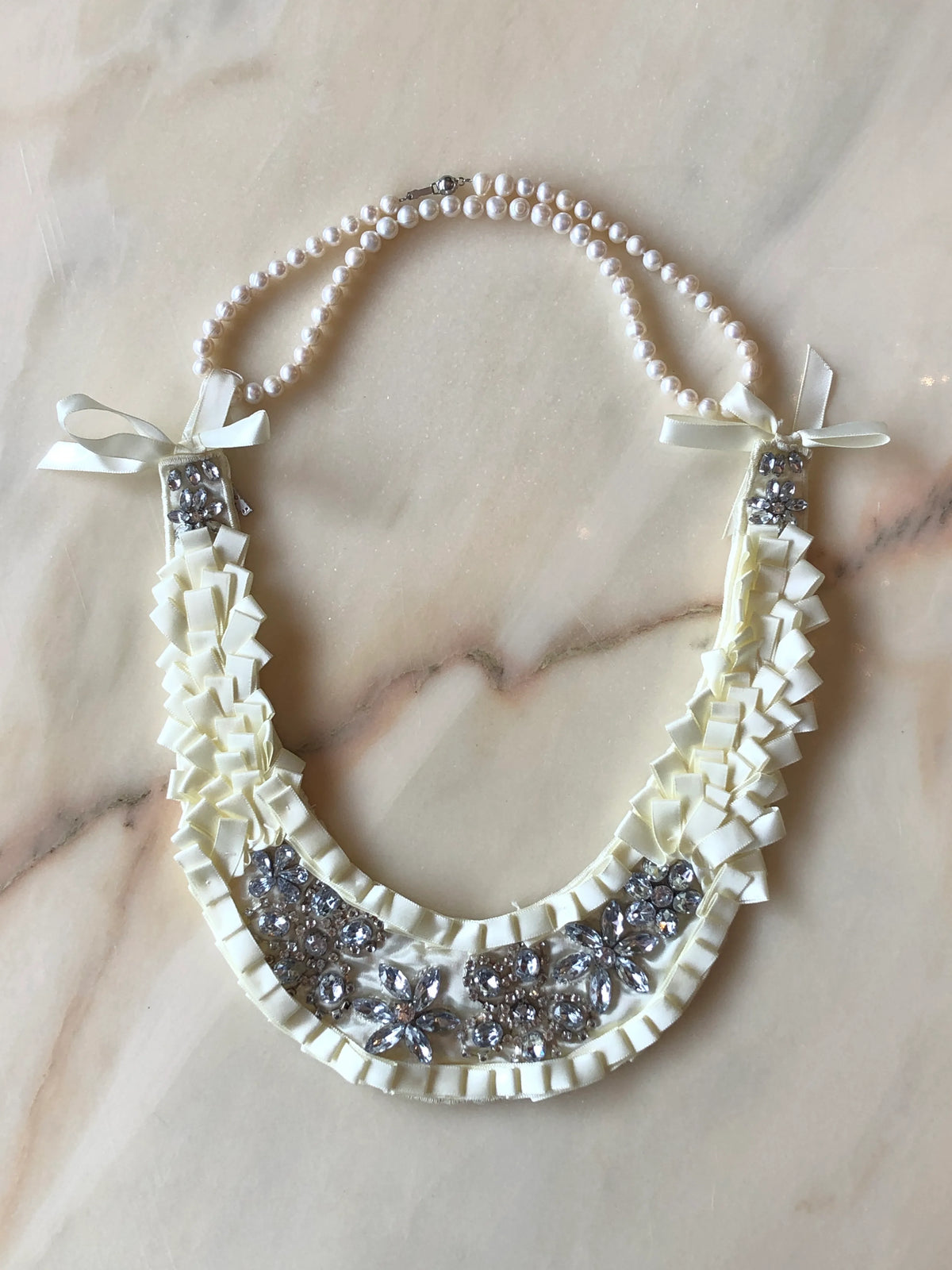 Broochella Maison Broche La Lumière Blanche ivory necklace bib featuring pearl strands, sculptural ivory ribbon ruffles, bows and sparkling crystal embellishments, photographed on a pale white marble background.