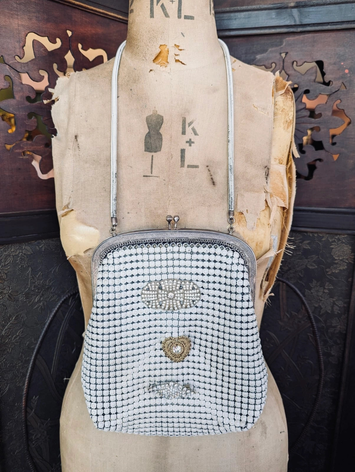 Broochella Maison Broche le bijou du soir vintage white glomesh bag, with vintage brooch embellishments. It is hung over a vintage mannequin   The background is a carved, dark wood screen.