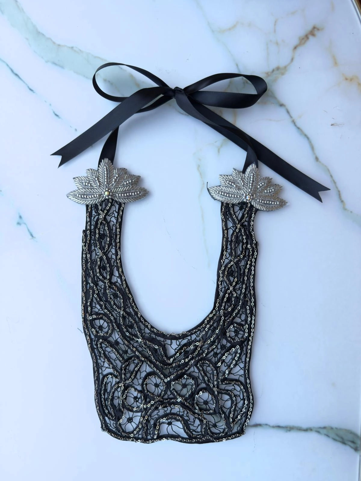 Broochella Maison Broche Le Coeur black, handmade lace and chain bib/necklace with silver embellishments and a ribbon tie. The background is white marble.