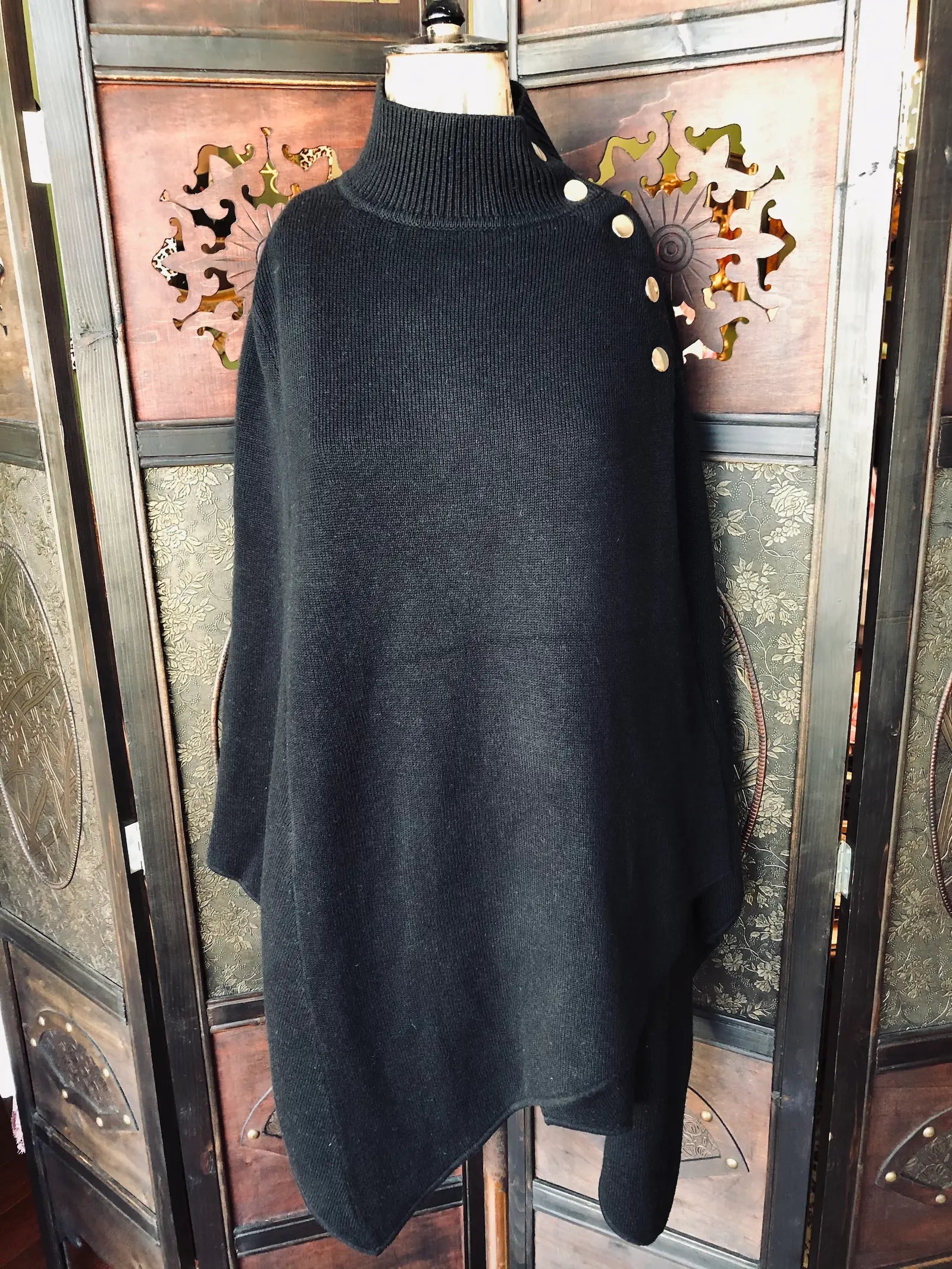 Broochella Maje, One Size, Black 47% Acrylic, 25% Viscose, 22% Wool, 5% Polyamide, 1% Elastane. High ribbed neckline. Gold-tone shoulder buttons. Asymmetrical hem. It is pictured on a vintage mannequin. The background is an ornate, vintage, dark wood room divider.