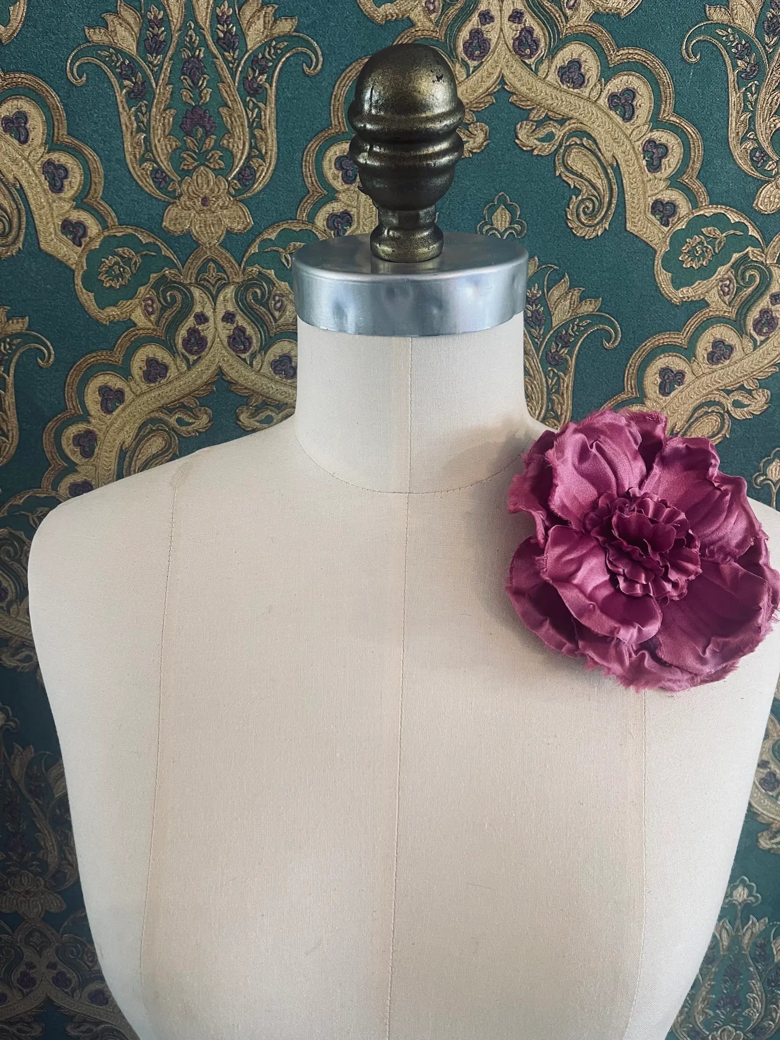 Broochella malva handmade fabric flower brooch in lilac. Styled on a mannequin. The background is green and gold damask wallpaper