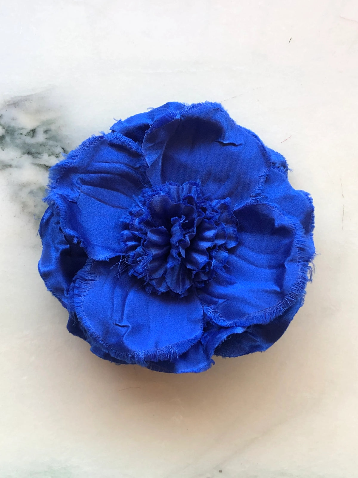 Broochella Malva Flower Brooch Pin in vivid cobalt blue, featuring layered fabric petals with softly frayed edges and a full ruffled floral centre on a cream marble background.