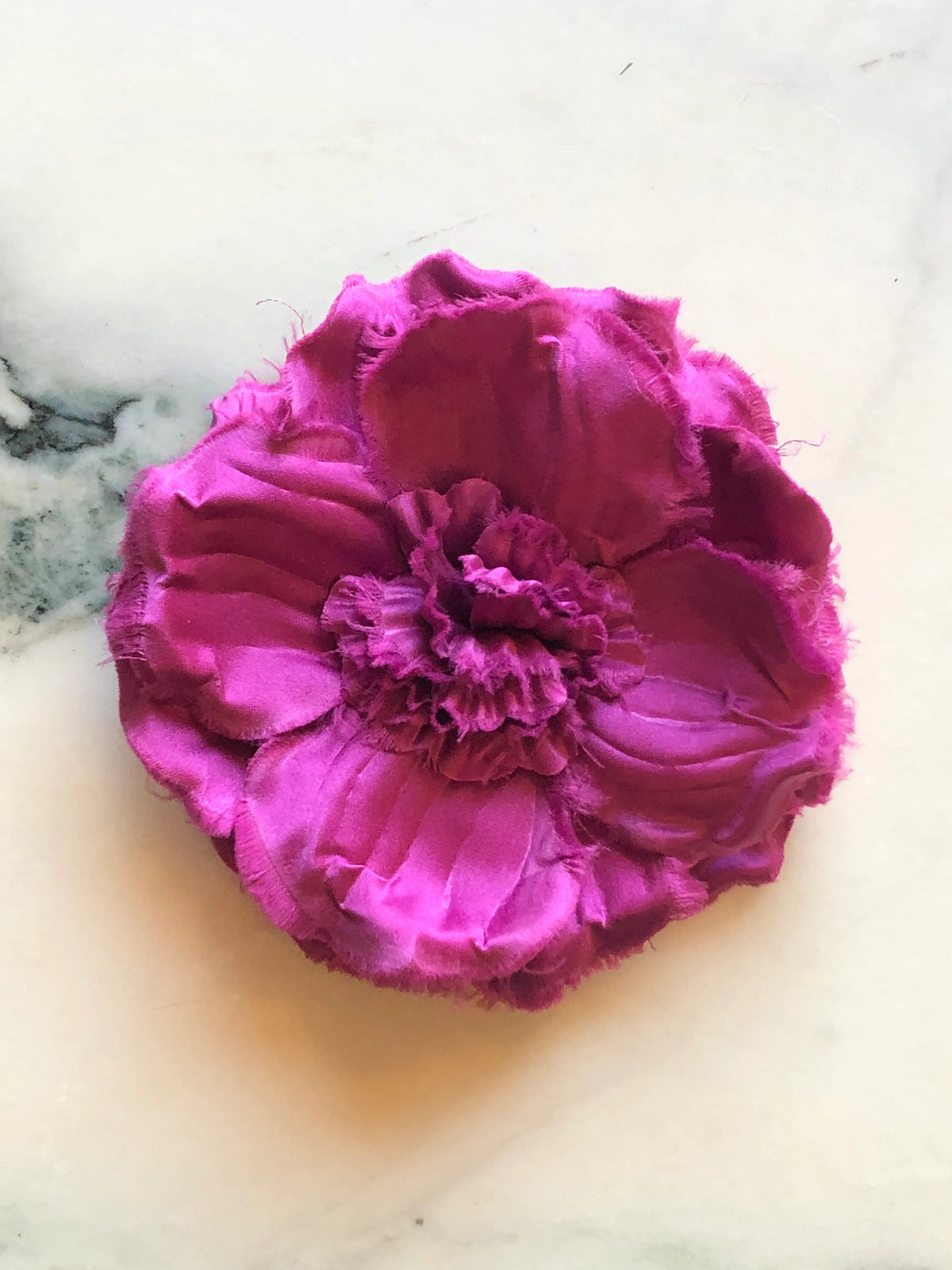 Broochella Malva Flower Brooch Pin in vibrant lilac pink, featuring layered fabric petals with softly frayed edges and a full ruffled floral centre on a cream marble background.