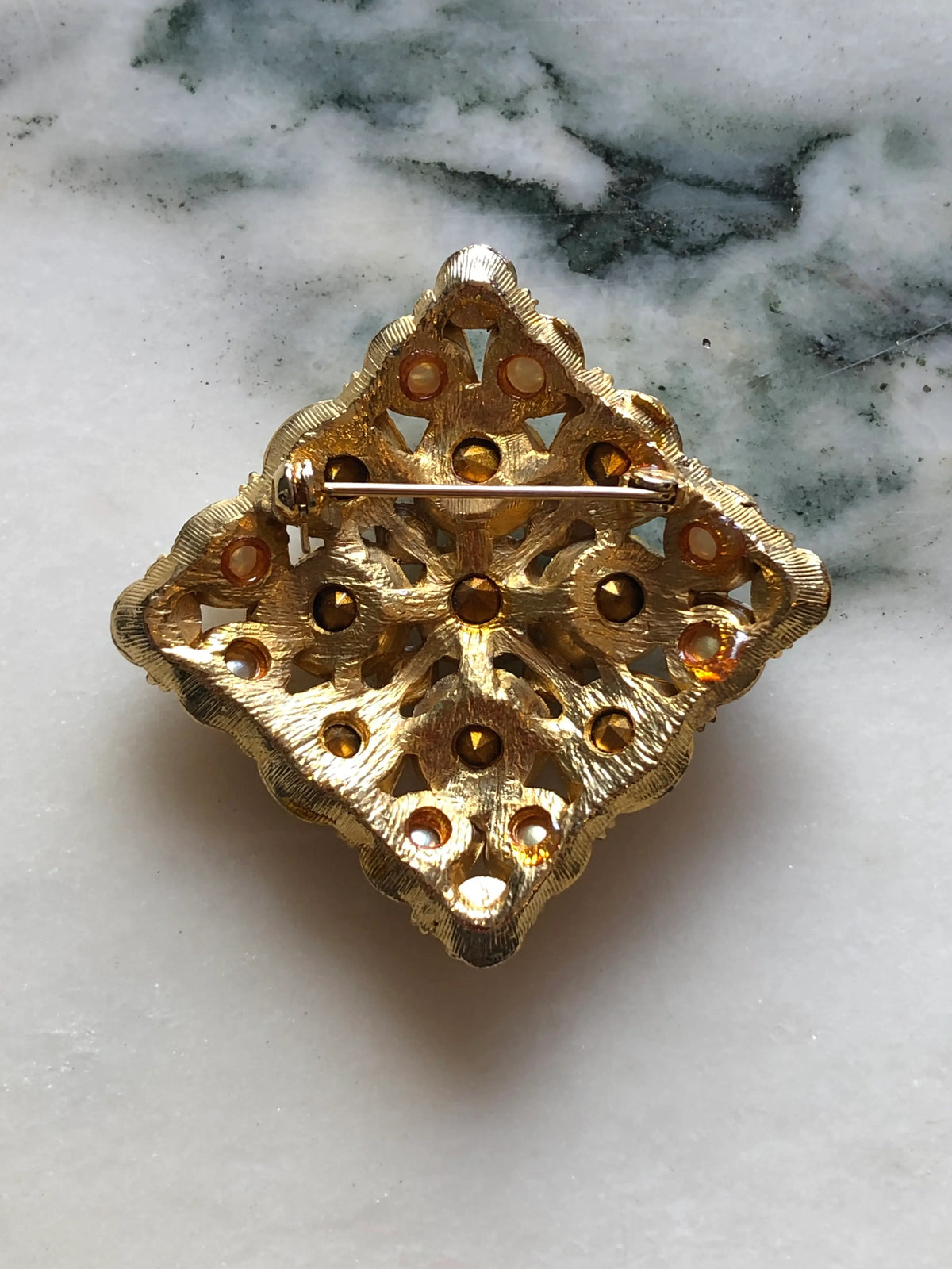 The back view of Broochella Marcella Vintage Pearl & Crystal Brooch featuring large clear faceted crystals and luminous faux pearls in an ornate gold-tone diamond-shaped setting on a cream and grey marble background.