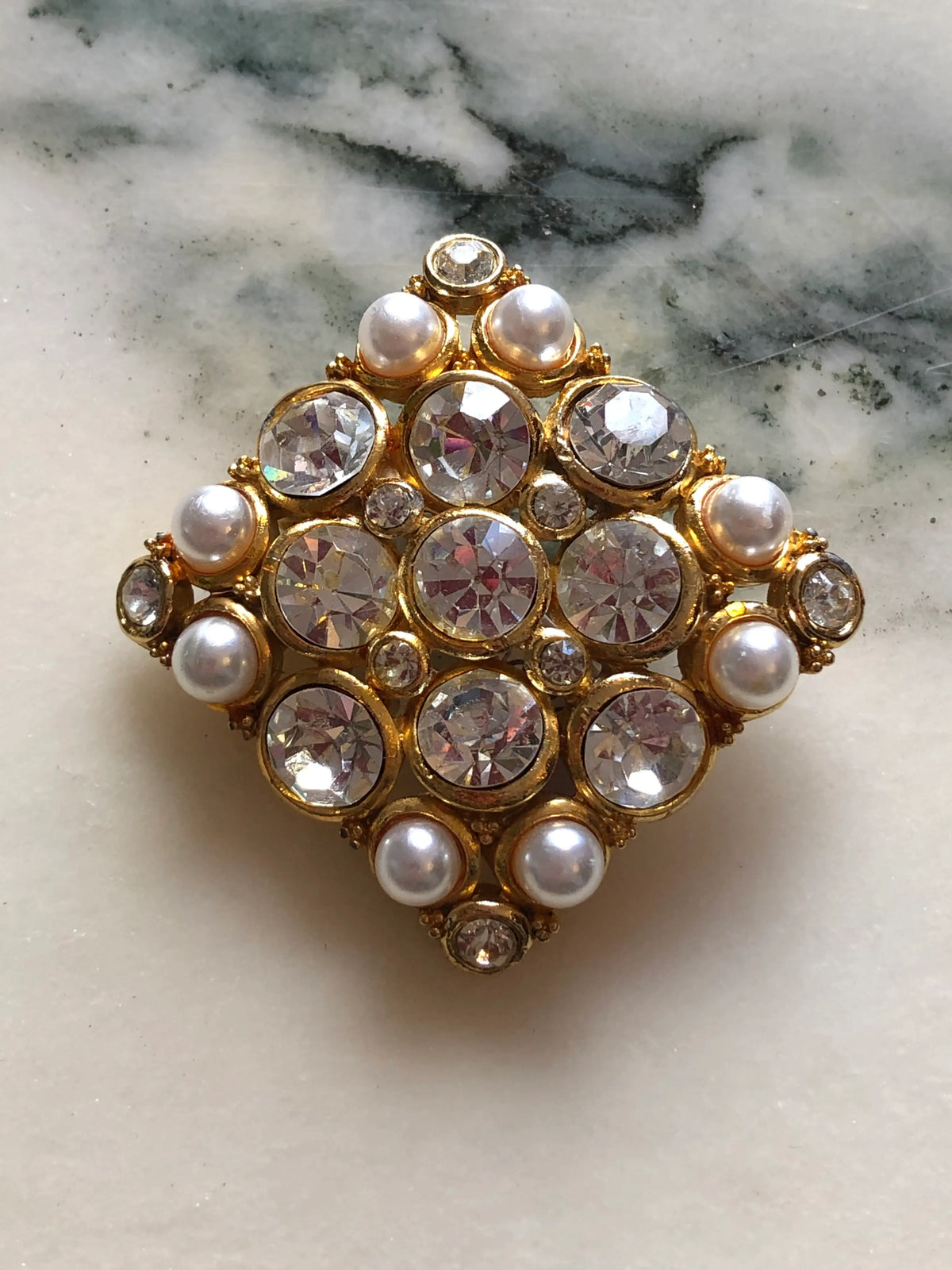 Broochella Marcella Vintage Pearl & Crystal Brooch featuring large clear faceted crystals and luminous faux pearls in an ornate gold-tone diamond-shaped setting on a cream and grey marble background.