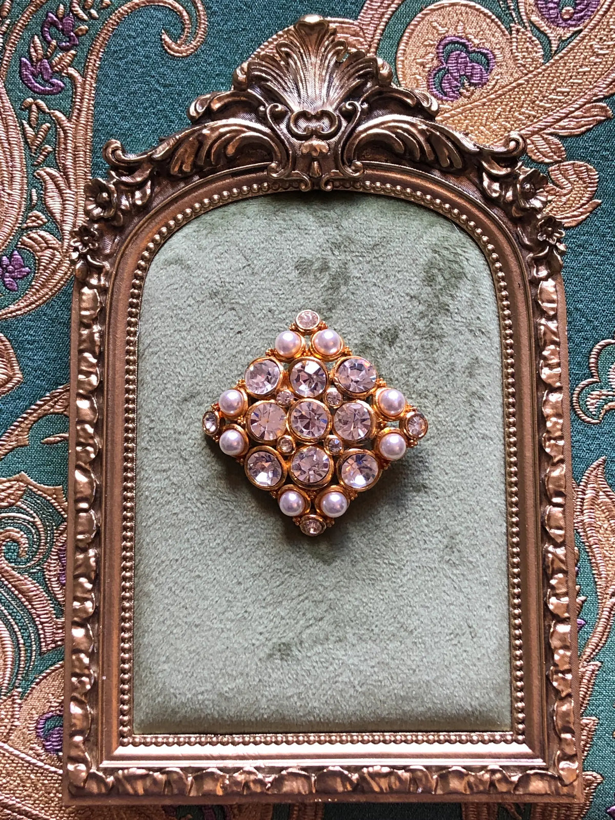 Broochella Marcella Vintage Pearl & Crystal Brooch featuring large clear faceted crystals and luminous faux pearls in an ornate gold-tone diamond-shaped setting. Pictured on a green velvet tray with an ornate gold frame. The background is green and gold damask wallpaper