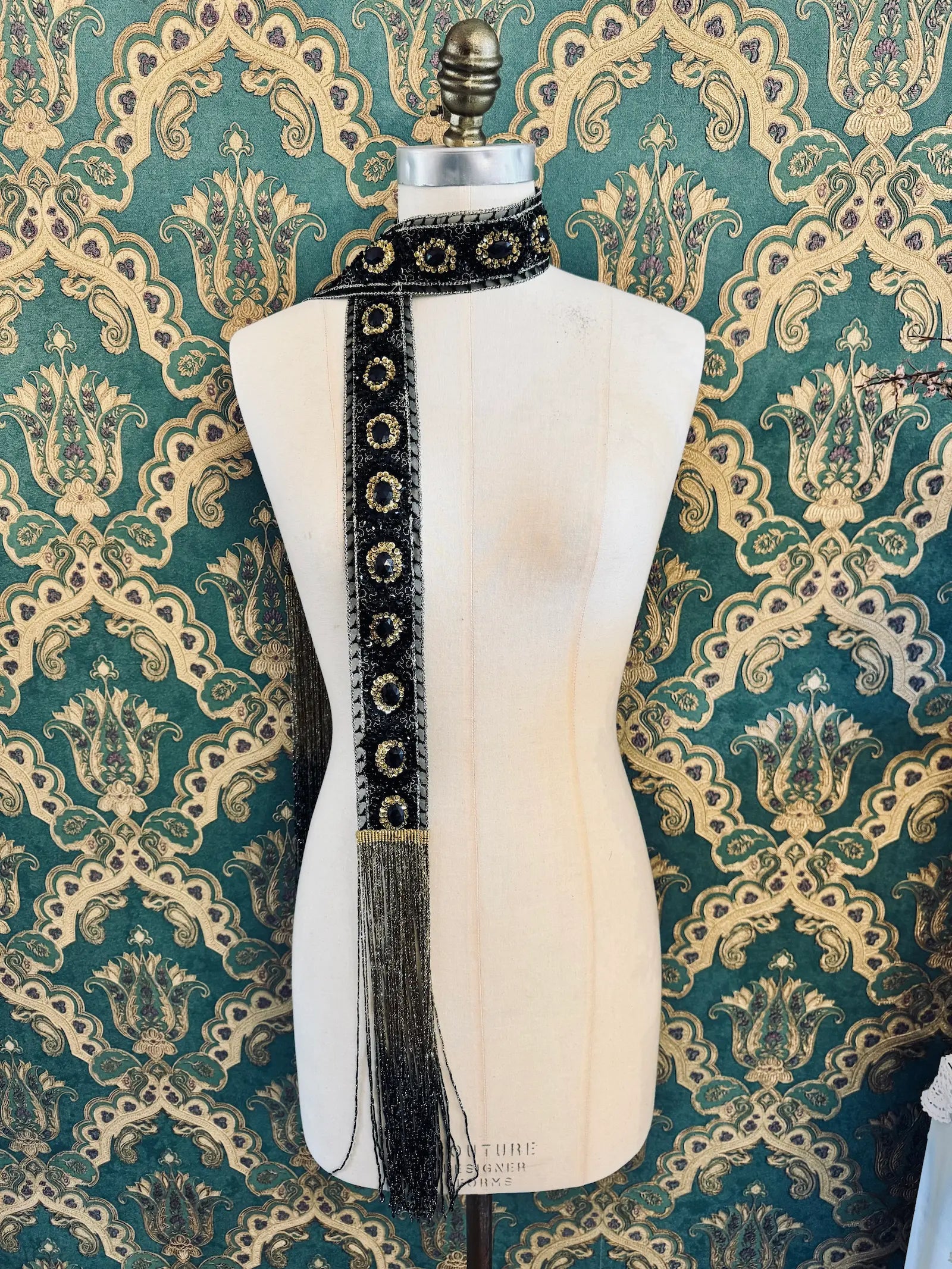 Broochella Antionette handmade fringe black and gold embellished foulard neck tie. Pictured on a mannequin. The background is green and gold damask wallpaper.