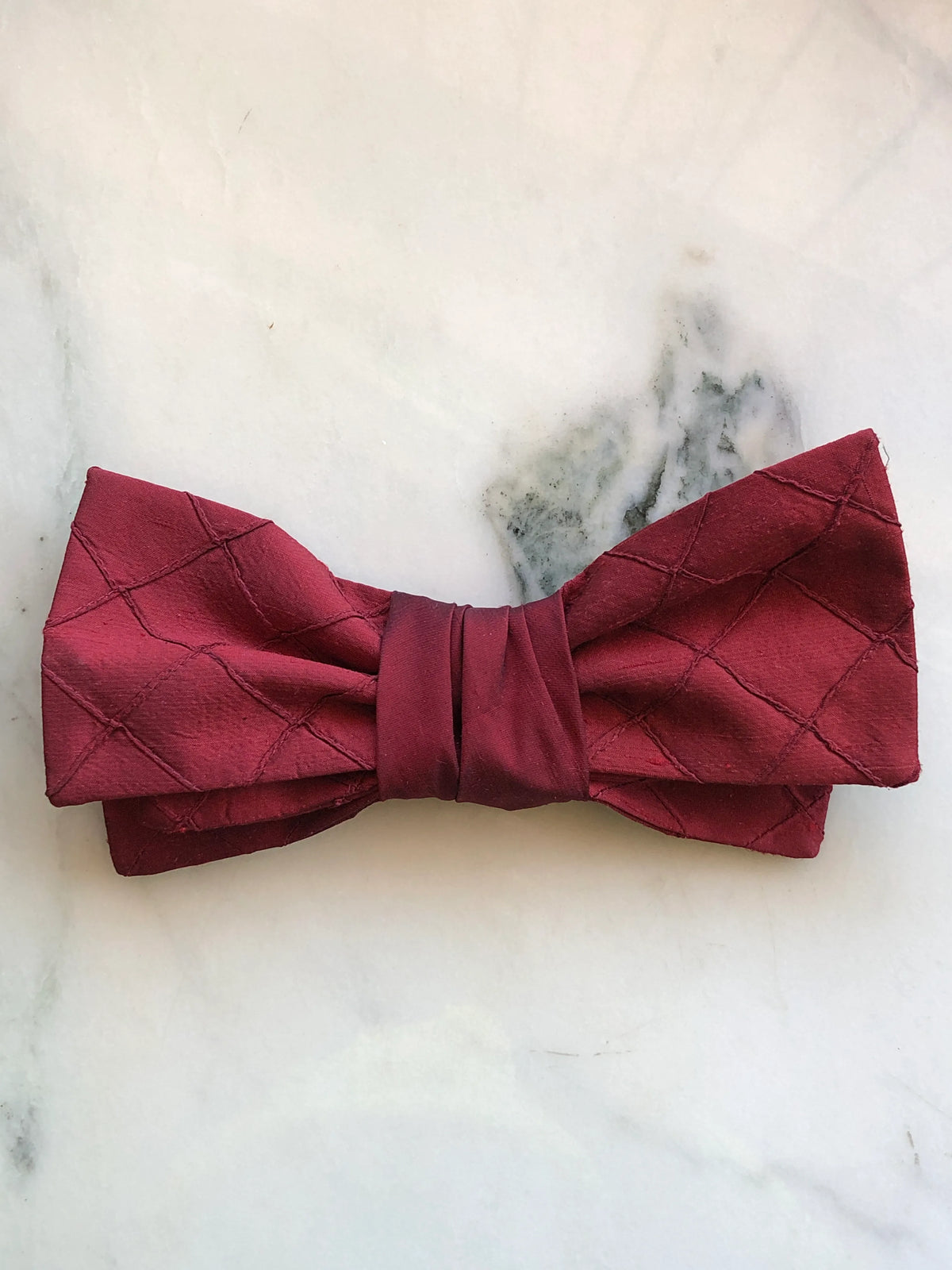 Broochella Marie Antoinette burgundy silk bow brooch pin in rich wine burgundy with a softly structured double bow and gathered wrapped centre, photographed on a white marble background with soft grey-green veining.