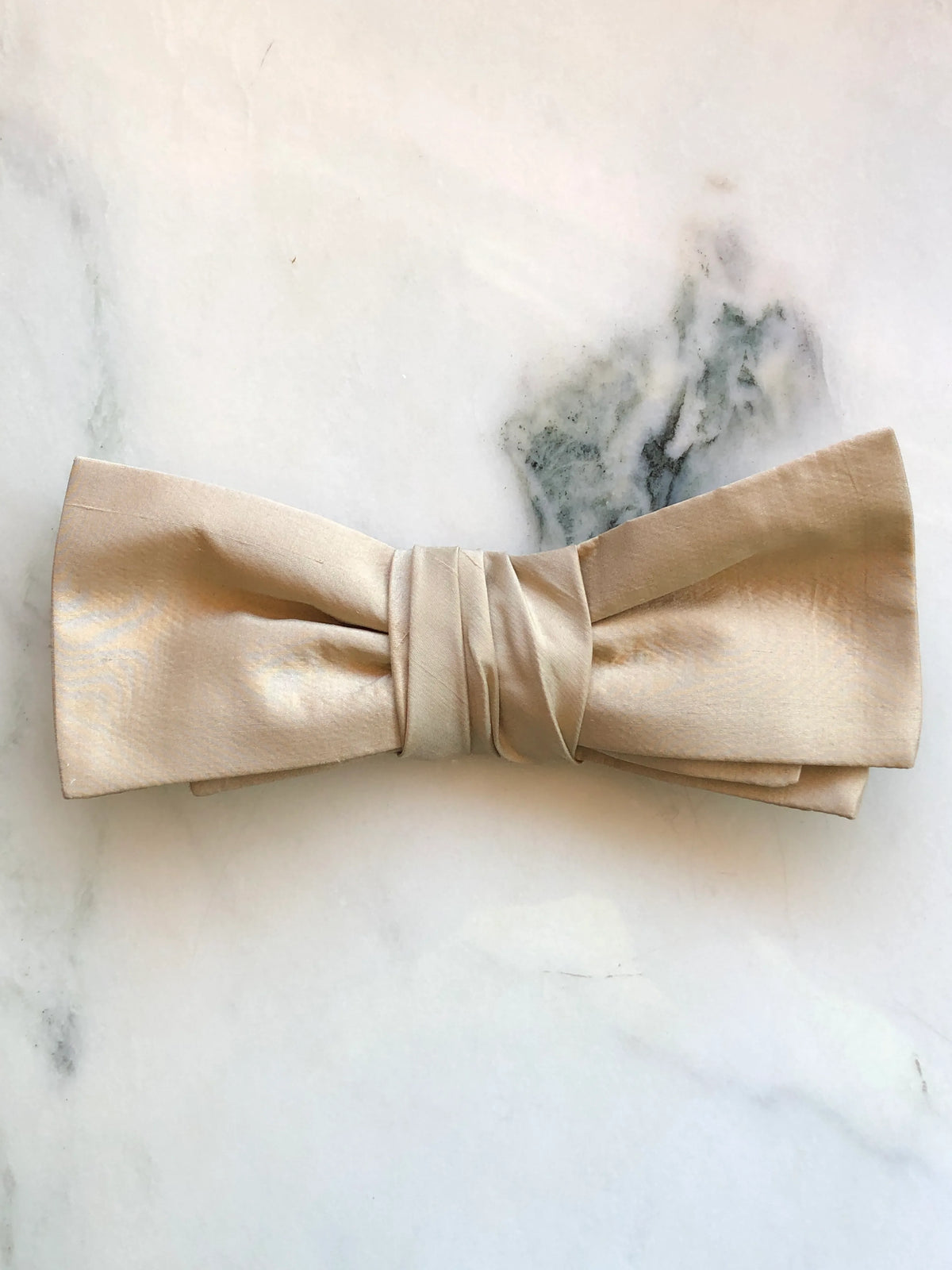 Broochella Marie Antoinette gold silk bow brooch pin in soft champagne gold silk with a structured double bow and gathered wrapped centre, photographed on a white marble background with soft grey-green veining.