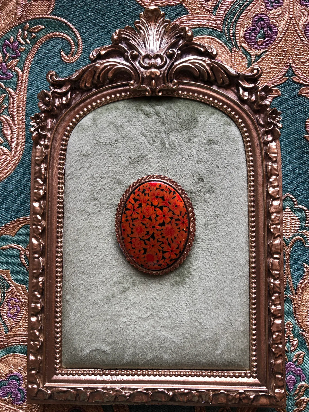 Broochella Marigold Garden vintage oval brooch with orange and amber flowers on a black background, a glossy domed centre and textured gold-tone frame, measuring approximately 4.5 × 3.5 cm. Photographed on a green velvet tray with an ornate gold frame. The background is green and gold damask wallpaper