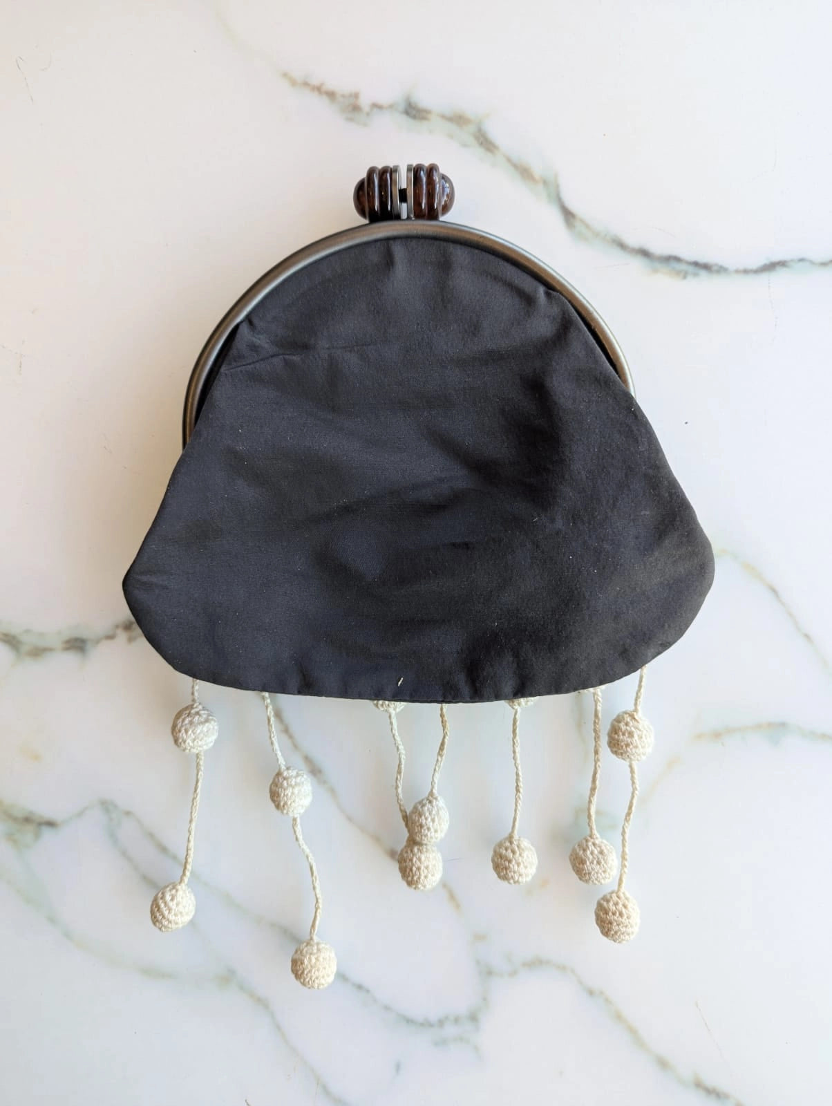 The back view of a A Broochella Marni grey cotton kiss-lock clutch purse with cream-coloured crochet fringe beads hanging from the clasp, displayed on a white marble surface.