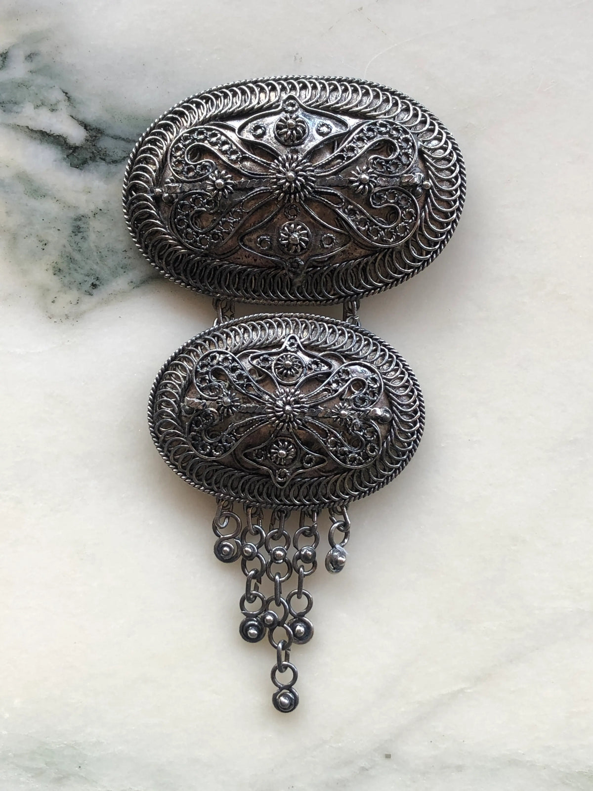 Broochella Medaglione Cascata Vintage Large Brooch Pin featuring two ornate silver-tone oval medallions with intricate filigree detailing and dangling metal charms, displayed on sage green velvet in an ornate antique gold frame against green and gold patterned fabric.