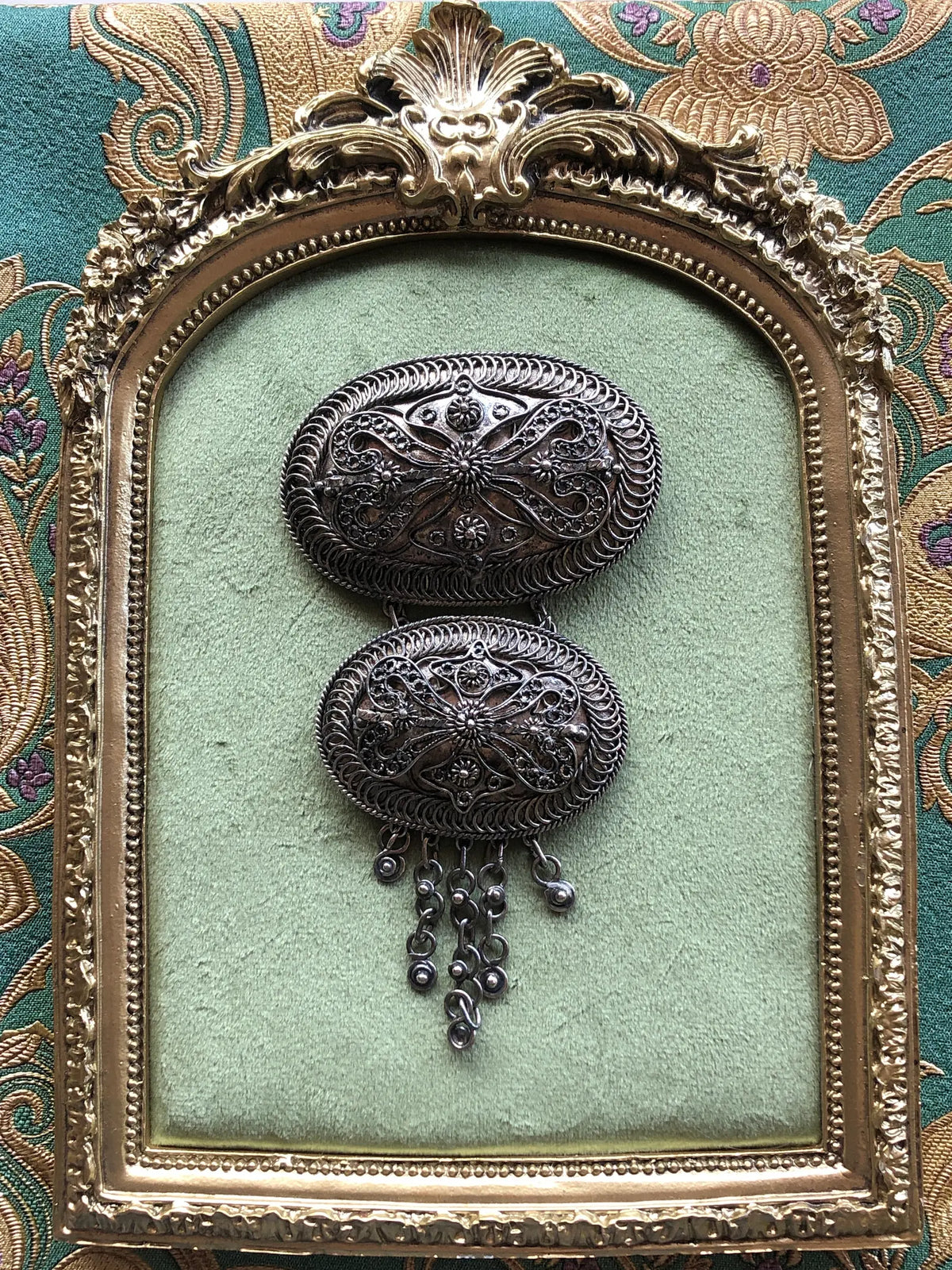 Broochella Medaglione Cascata Vintage Large Brooch Pin featuring two ornate silver-tone oval medallions with intricate filigree detailing and dangling metal charms, displayed on sage green velvet in an ornate antique gold frame against green and gold patterned fabric.