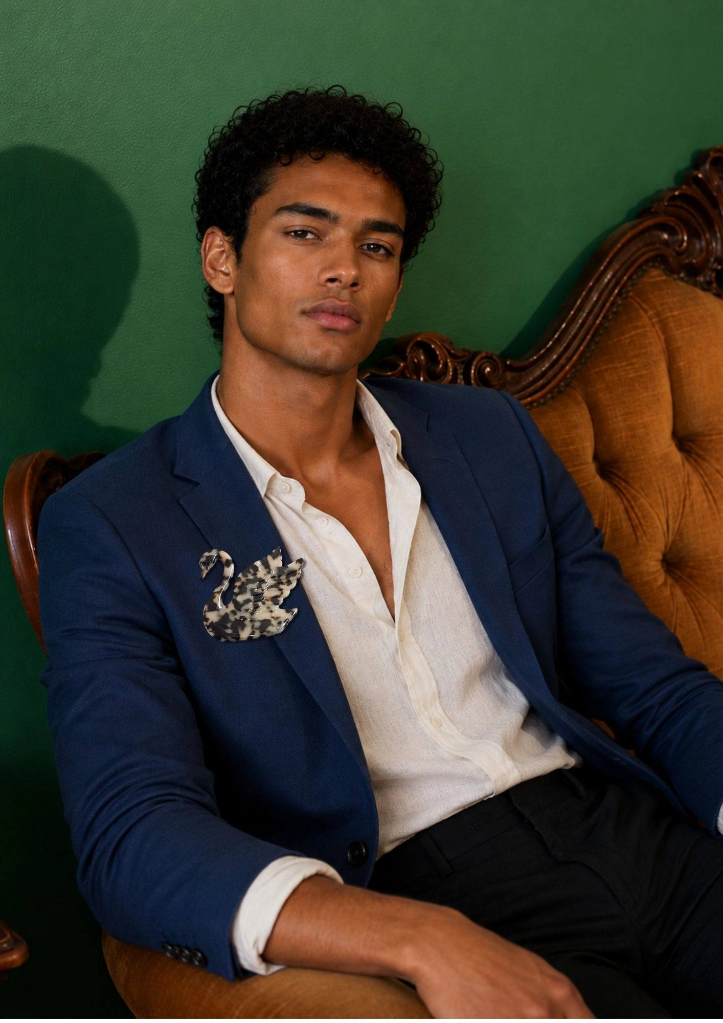 A man wearing a navy blue blazer with a decorative Broochella Men's Animalia tortoiseshell swan brooch on the chest, worn open over a white shirt, seated on an ornate tufted brown chair against a dark green wall.