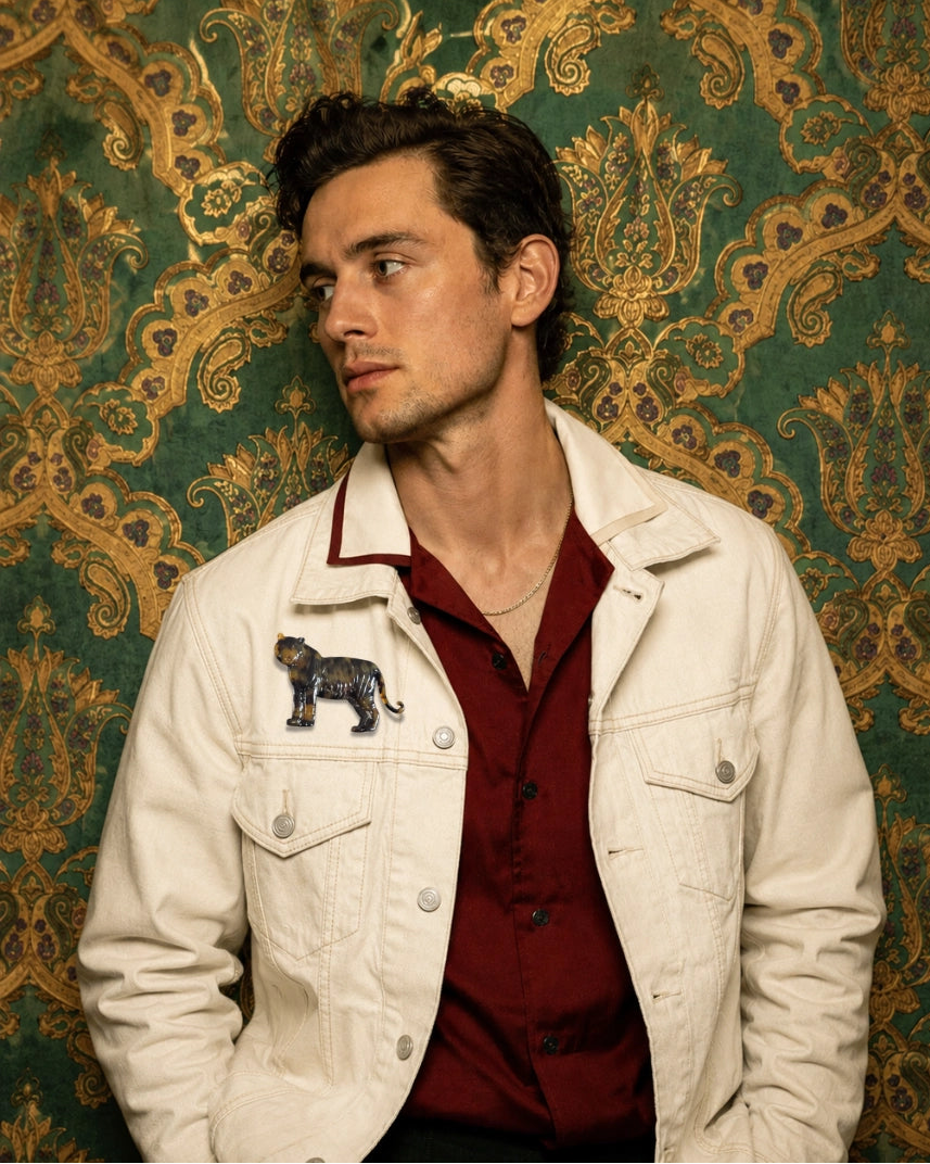 A man wearing a cream denim jacket with a Broochella Men's Animalia tortoiseshell tiger brooch on the chest, layered over a dark red button-down shirt, posed against a green and gold damask patterned backdrop.