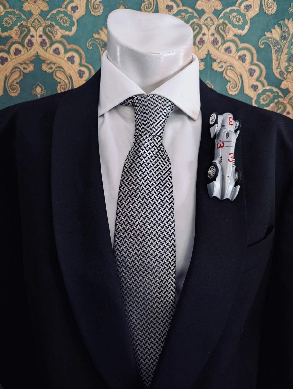 Broochella men's scuderia d'oro argento number three silver, vintage formula one racing car brooch. Pinned on a men's mannequin wearing a black suit and a houndstooth tie. The background is green and gold damask wallpaper. 