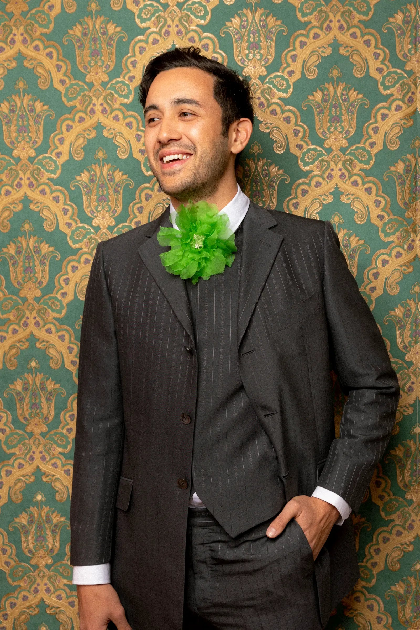 A man wearing a dark gray pinstripe three-piece suit with a bright green Broochella men's flower brooch, standing against a green and gold damask patterned wallpaper background.