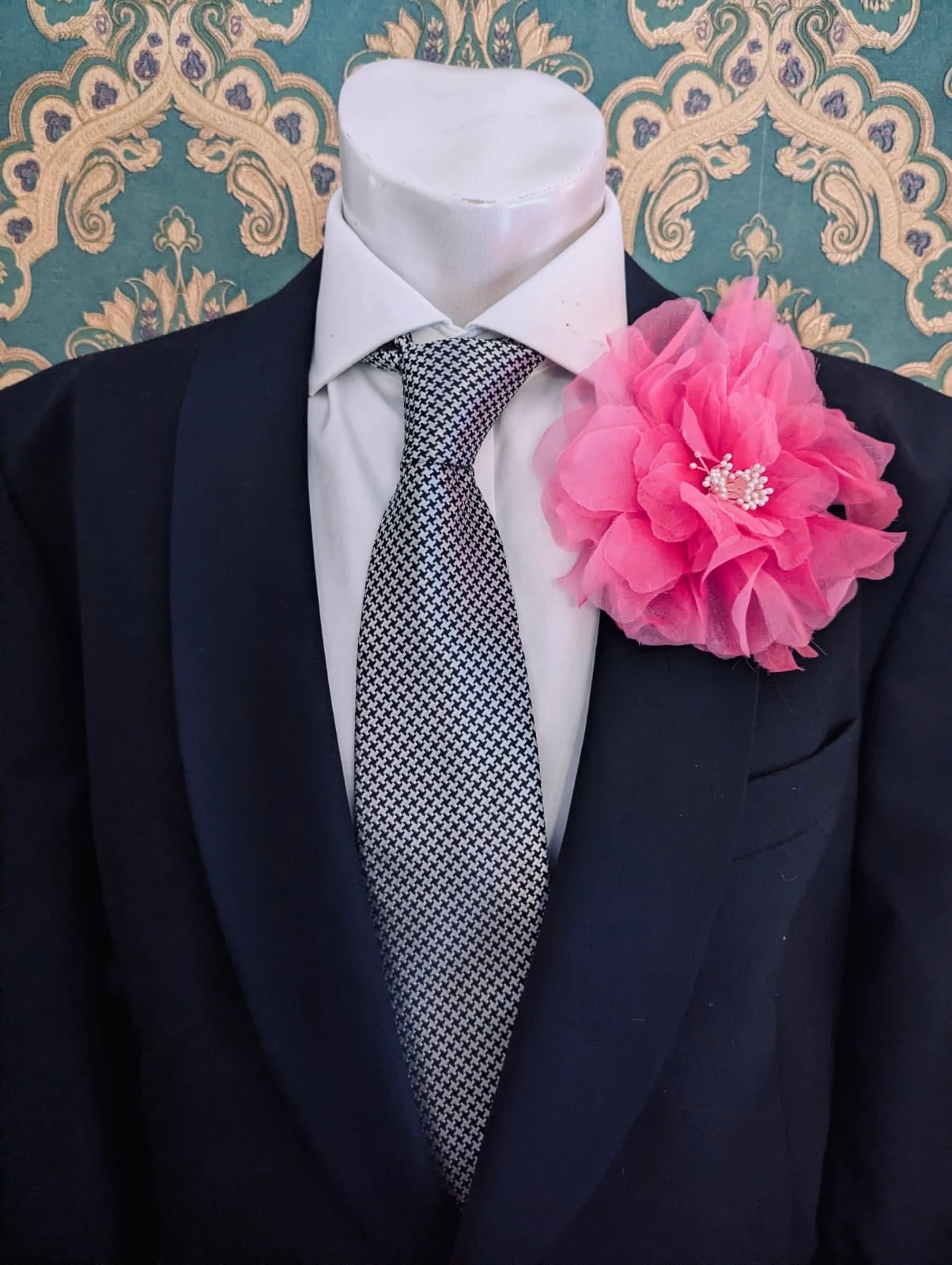 A black suit jacket displayed on a white mannequin torso, styled with a white dress shirt, a patterned grey and white tie, and a Broochella men's pink  Aubrieta flower brooch against a teal and gold damask wallpaper background.