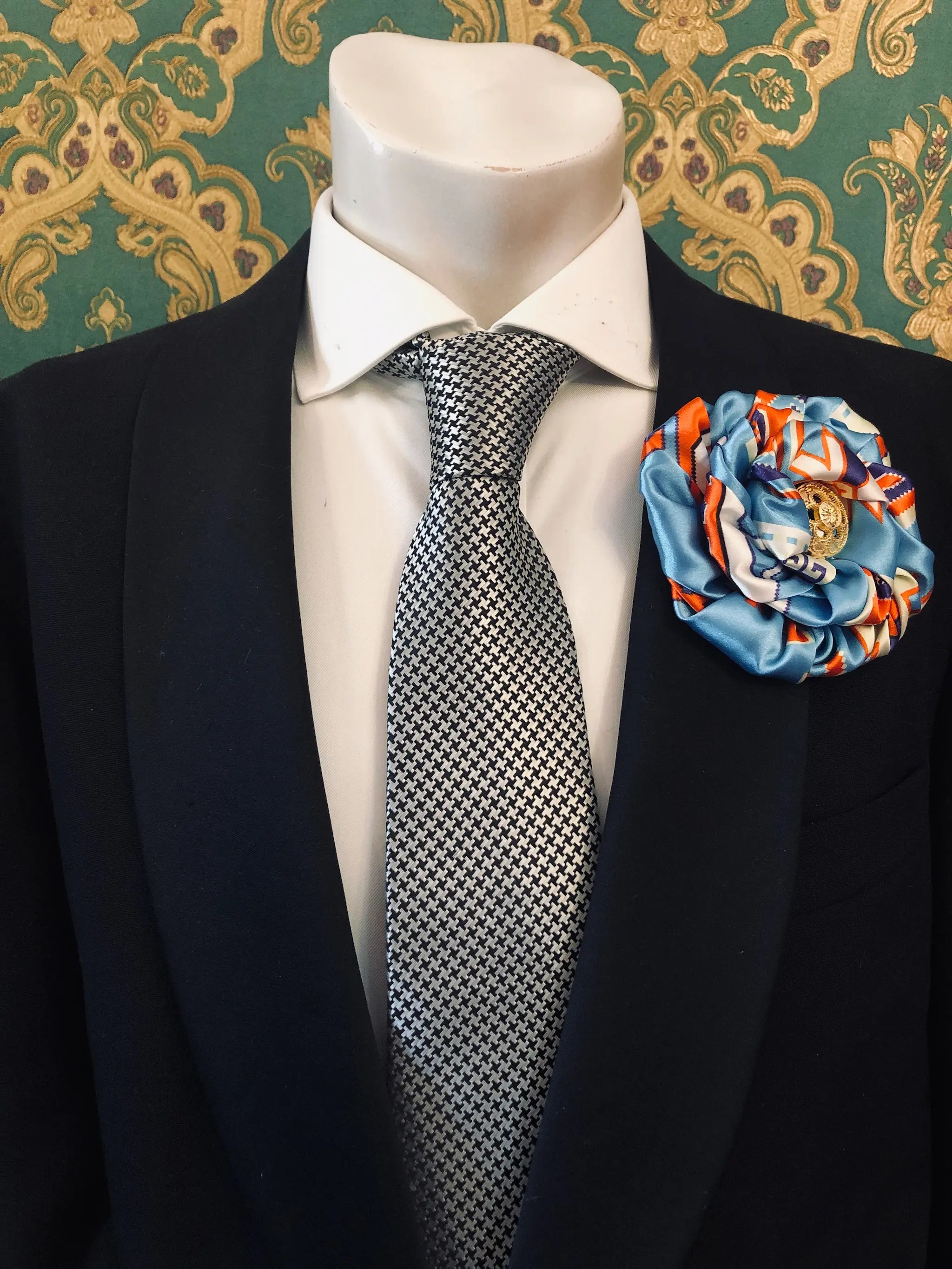 A men's formal outfit displayed on a white mannequin torso, featuring a black suit jacket, a white dress shirt, a black-and-white patterned tie, and a large Broochella Men's Bougainville decorative fabric flower brooch in light blue, orange, and gold on the lapel. The background is ornate green and gold patterned wallpaper.