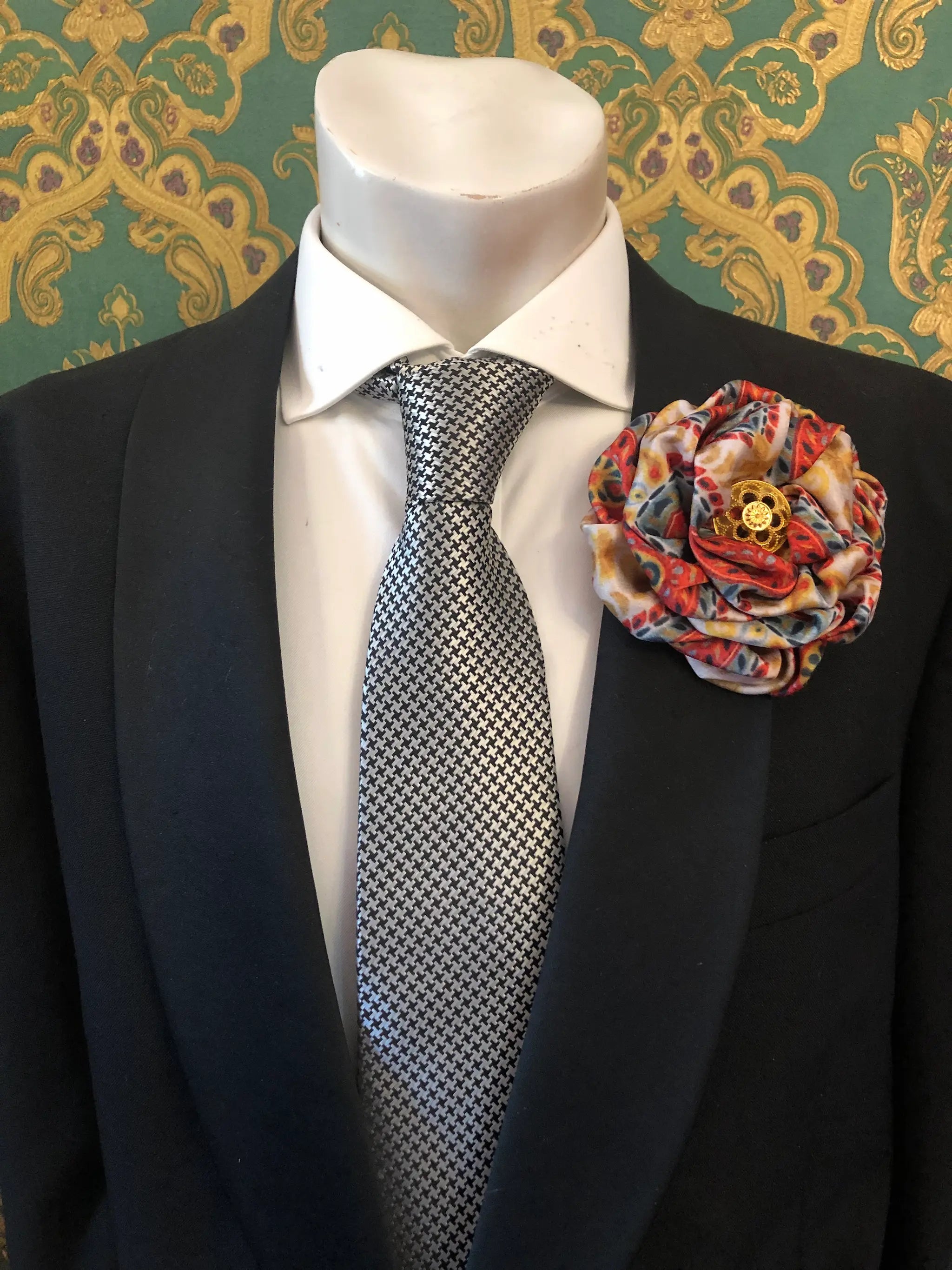 A men's formal outfit displayed on a white mannequin torso, featuring a black suit jacket, a white dress shirt, a black-and-white patterned tie, and a large Broochella Men's Bougainville decorative fabric flower brooch in orange print on the lapel. The background is ornate green and gold patterned wallpaper.