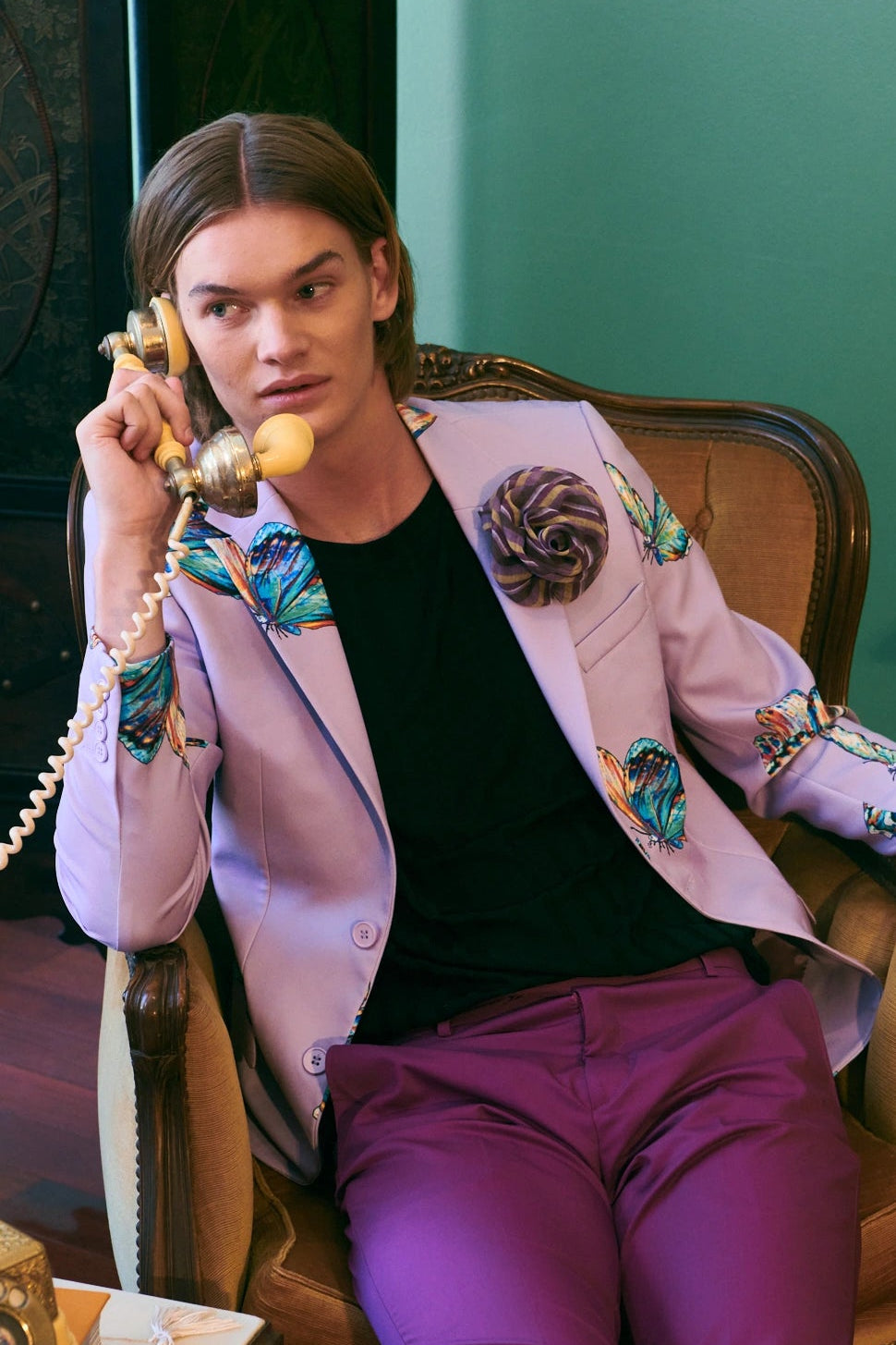 A model sits in a vintage leather armchair, holding a retro rotary telephone. The model is wearing a lavender blazer with colorful floral embroidery on the lapels and sleeves, a black t-shirt, and magenta trousers. He is wearing the Broochella men's brassica flower brooch in purple stripe. 
