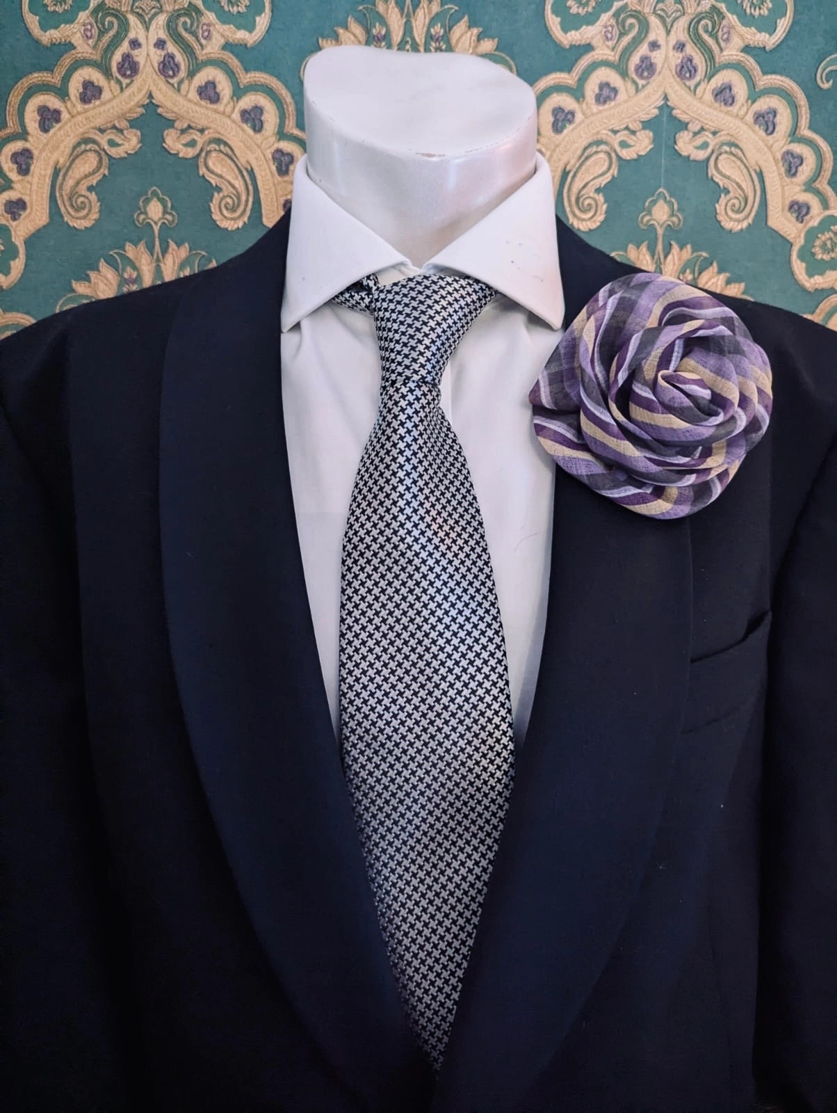 A men's formal outfit displayed on a white mannequin torso, featuring a dark navy suit jacket, a white dress shirt, a gray and white patterned tie, and the Broochella brassica men's flower brooch in green and purple stripes on the lapel, set against a teal and gold damask wallpaper background.