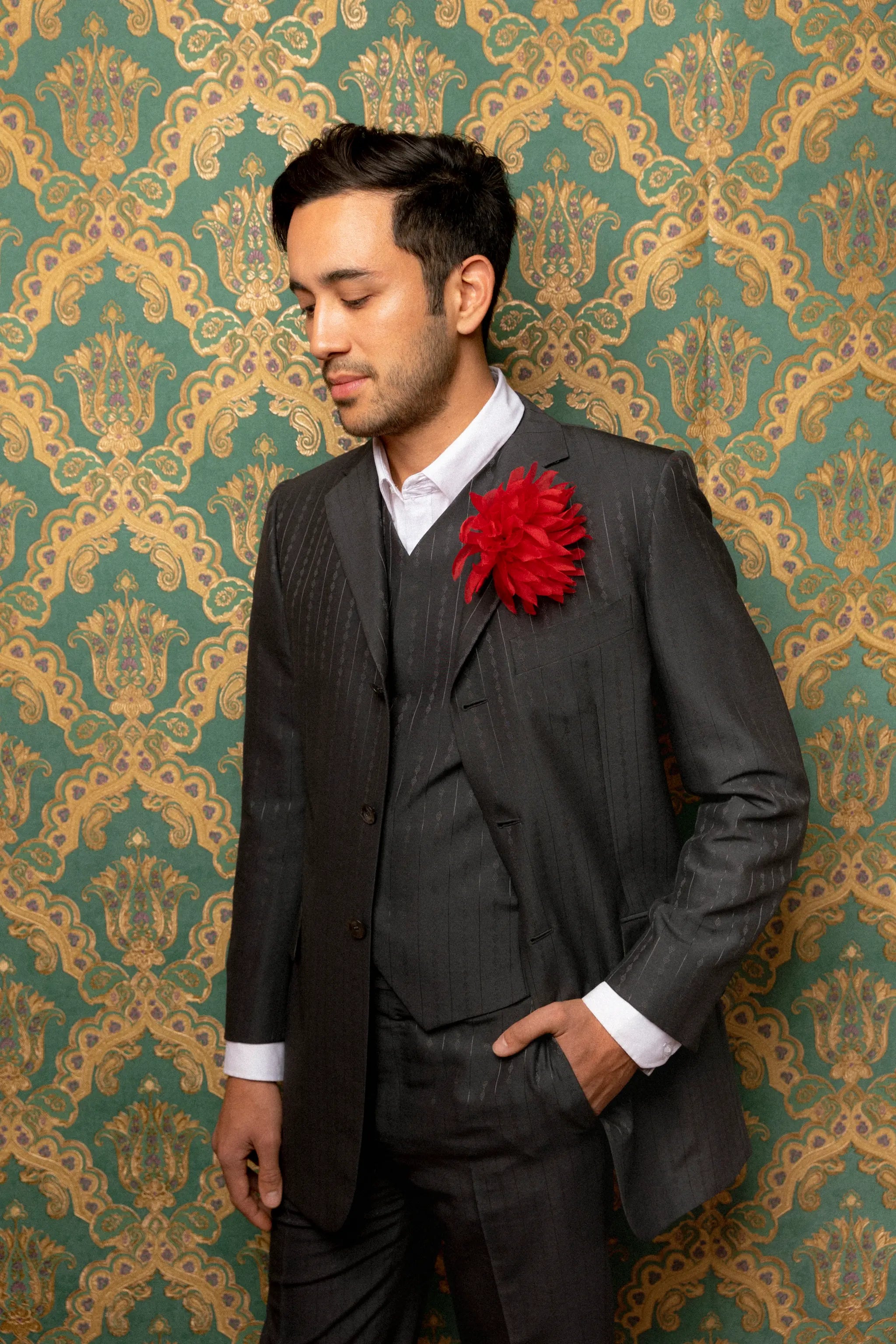 A man wearing a dark gray pinstripe three-piece suit with a white dress shirt and a bright red Broochella Dahlia flower brooch, standing against a green and gold damask patterned wallpaper background.