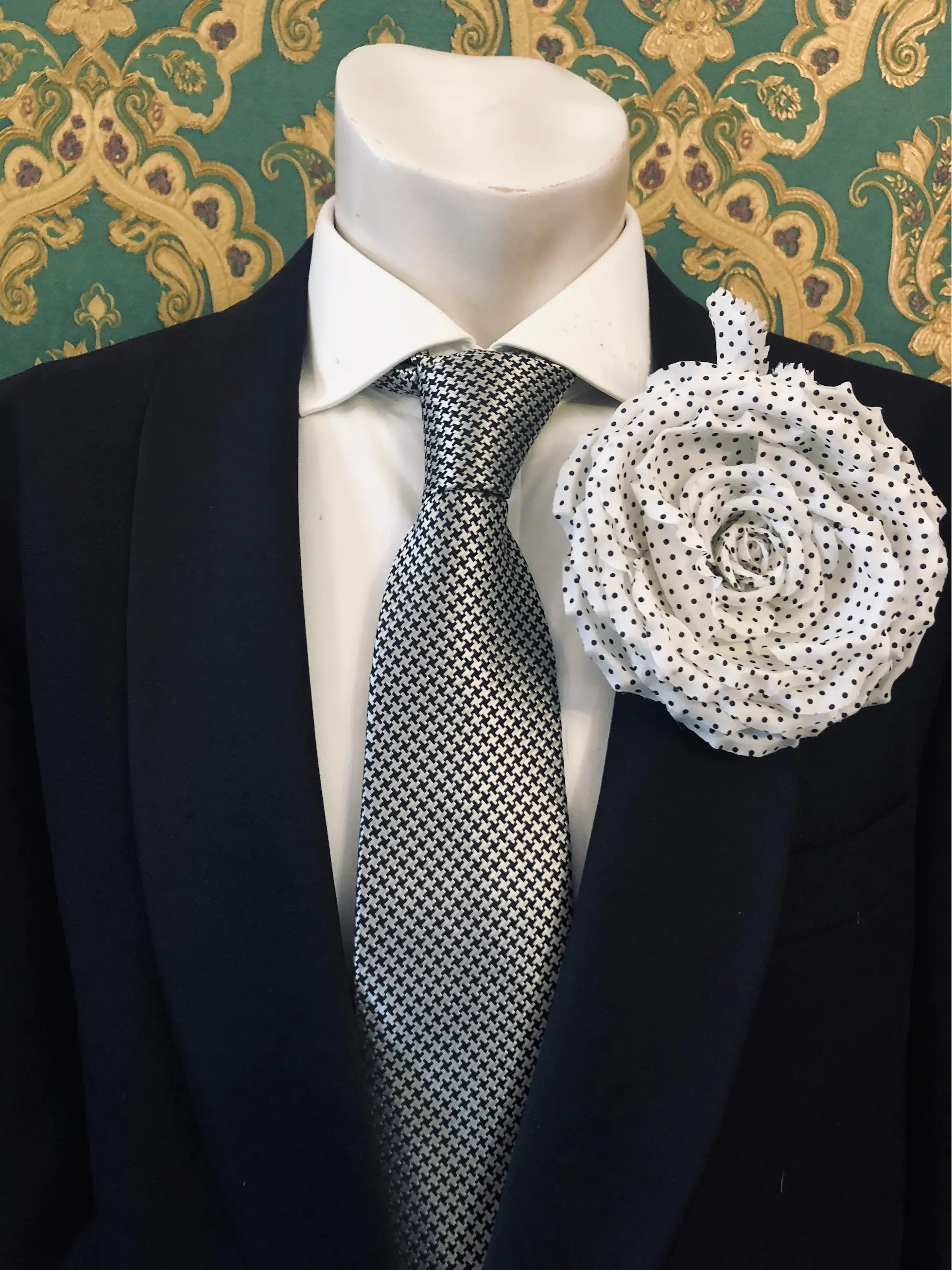 A Broochella Men's Dalmata white fabric flower brooch with black polka dots pinned to the lapel of a black suit jacket, styled with a white dress shirt and a black-and-white checkered tie, set against a green and gold patterned wallpaper background.