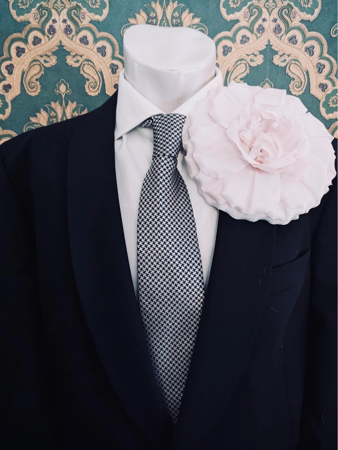 A formal outfit displayed on a white mannequin torso, featuring a black suit jacket, a white dress shirt, a grey and black patterned tie, and a large Broochella handmade men's femmina pale pink fabric flower brooch on the left lapel. The background is a teal and beige damask patterned wall.