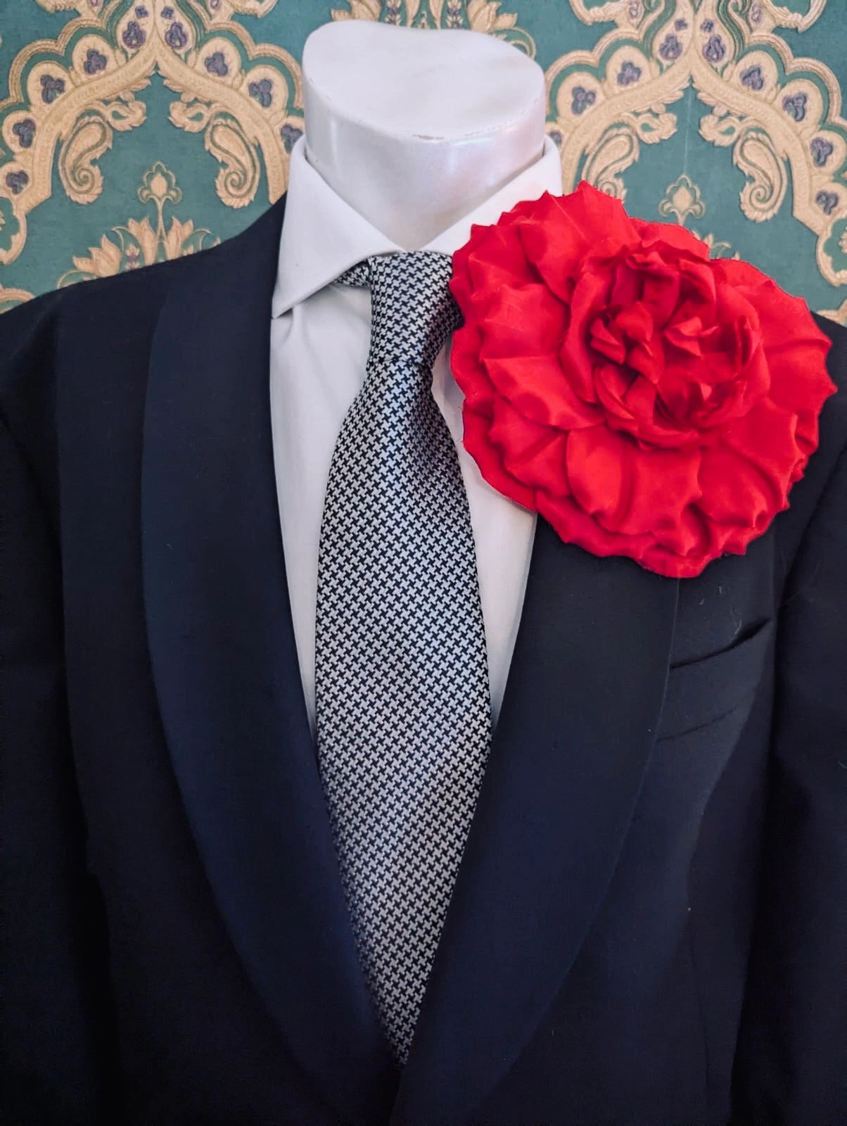 A formal outfit displayed on a white mannequin torso, featuring a black suit jacket, a white dress shirt, a grey and black patterned tie, and a large Broochella handmade men's femmina red fabric flower brooch on the left lapel. The background is a teal and beige damask patterned wall.