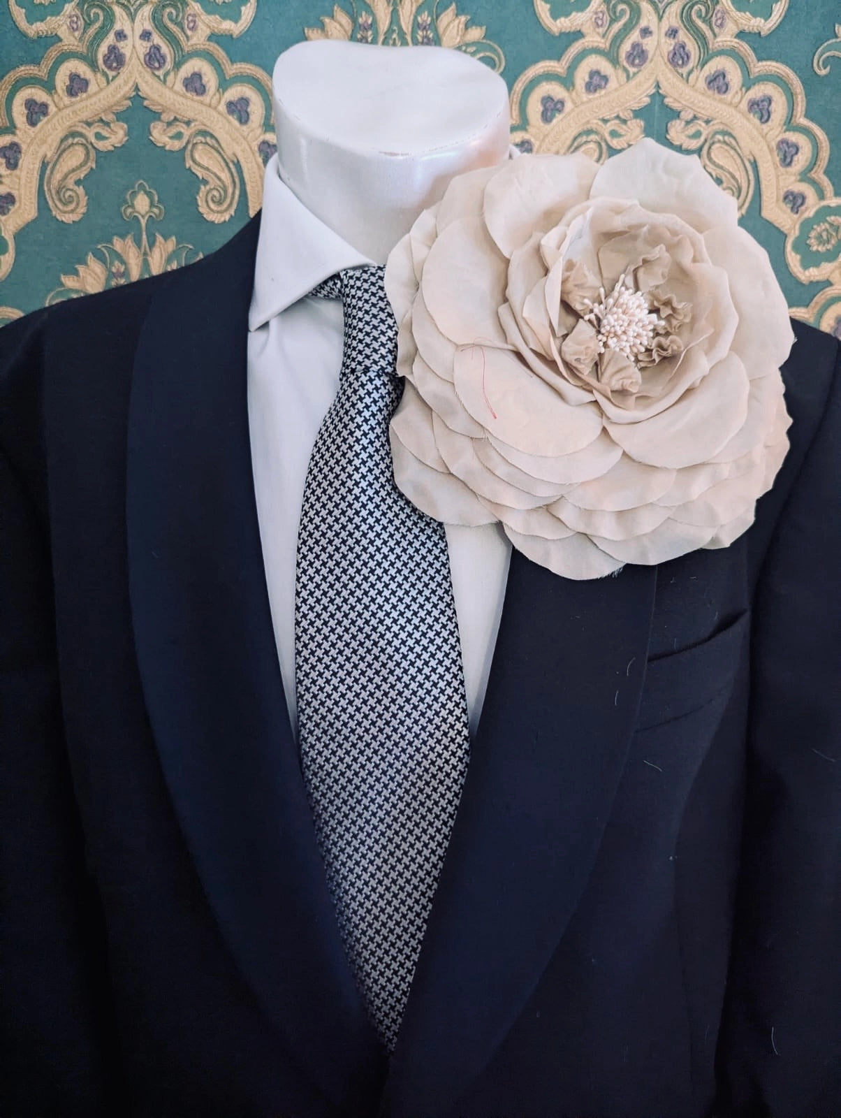 A large, Broochella handmade men's fiorita fabric flower brooch in beige, pinned to the lapel of a black suit jacket over a white dress shirt and a small patterned tie, displayed on a mannequin torso against a teal and gold patterned wallpaper background.
