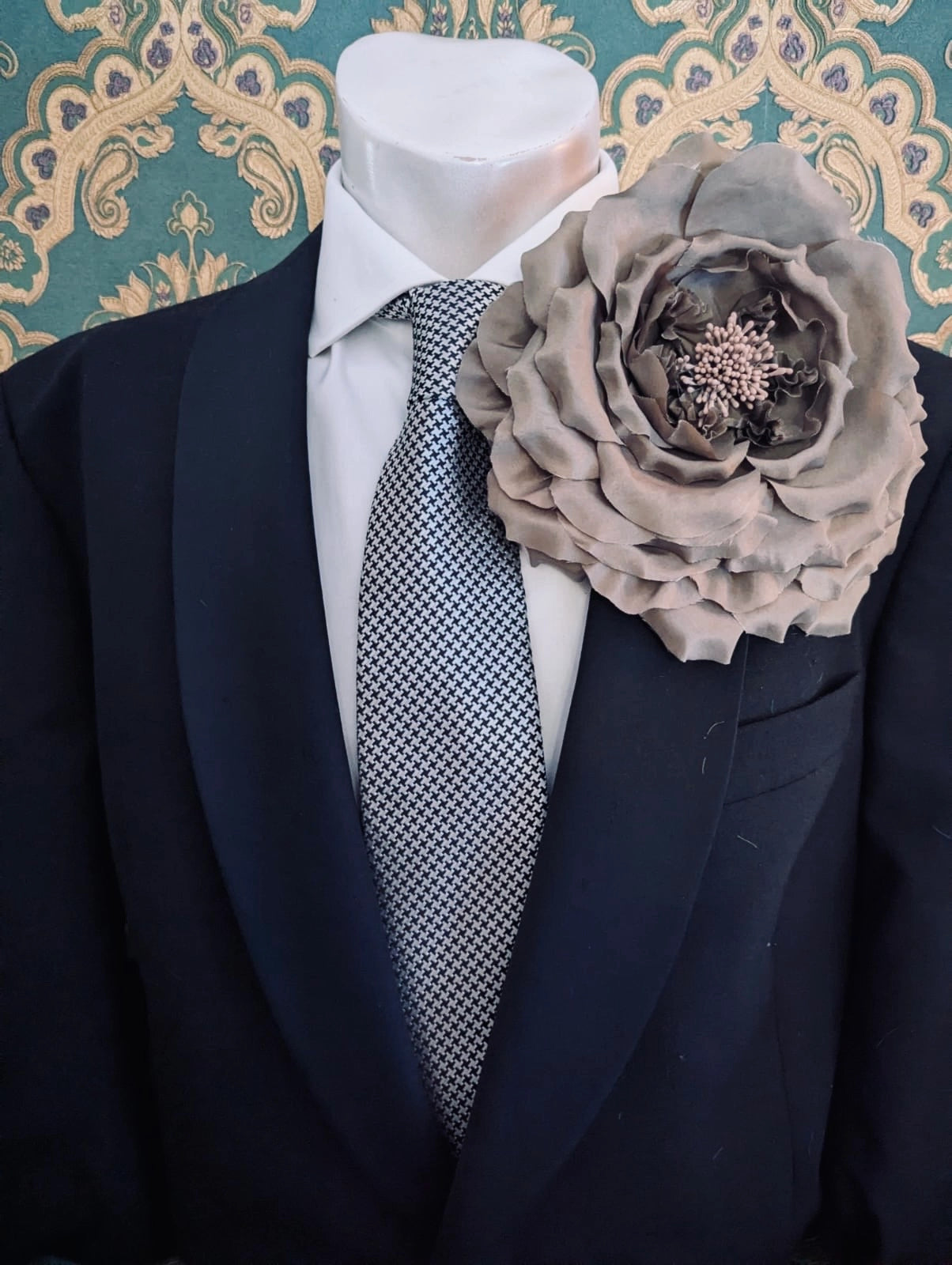 A large, Broochella handmade men's fiorita fabric flower brooch in brown, pinned to the lapel of a black suit jacket over a white dress shirt and a small patterned tie, displayed on a mannequin torso against a teal and gold patterned wallpaper background.