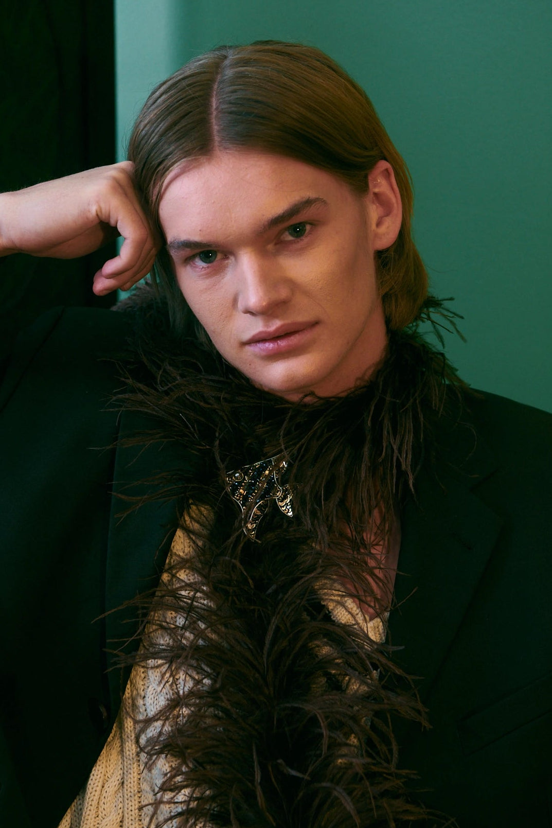 A model with long light brown hair poses in a black blazer and a dark brown feather boa over a beige knit sweater, set against a dark green wall. He is wearing the Broochella men's hand beaded gold and black fish brooch. 