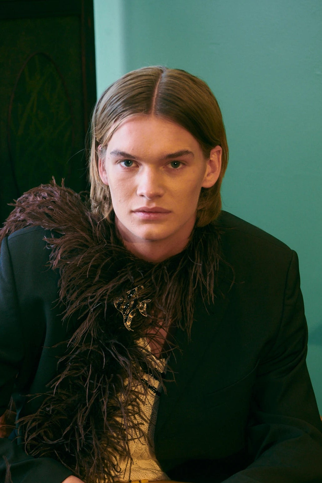 A man with long light brown hair sits in a brown leather armchair wearing a black jacket with dark brown feathered detailing over a cream shirt and bright yellow trousers. A matching yellow headrest with dark feathers rests on his lap. He is wearing the Broochella men's fish brooch.