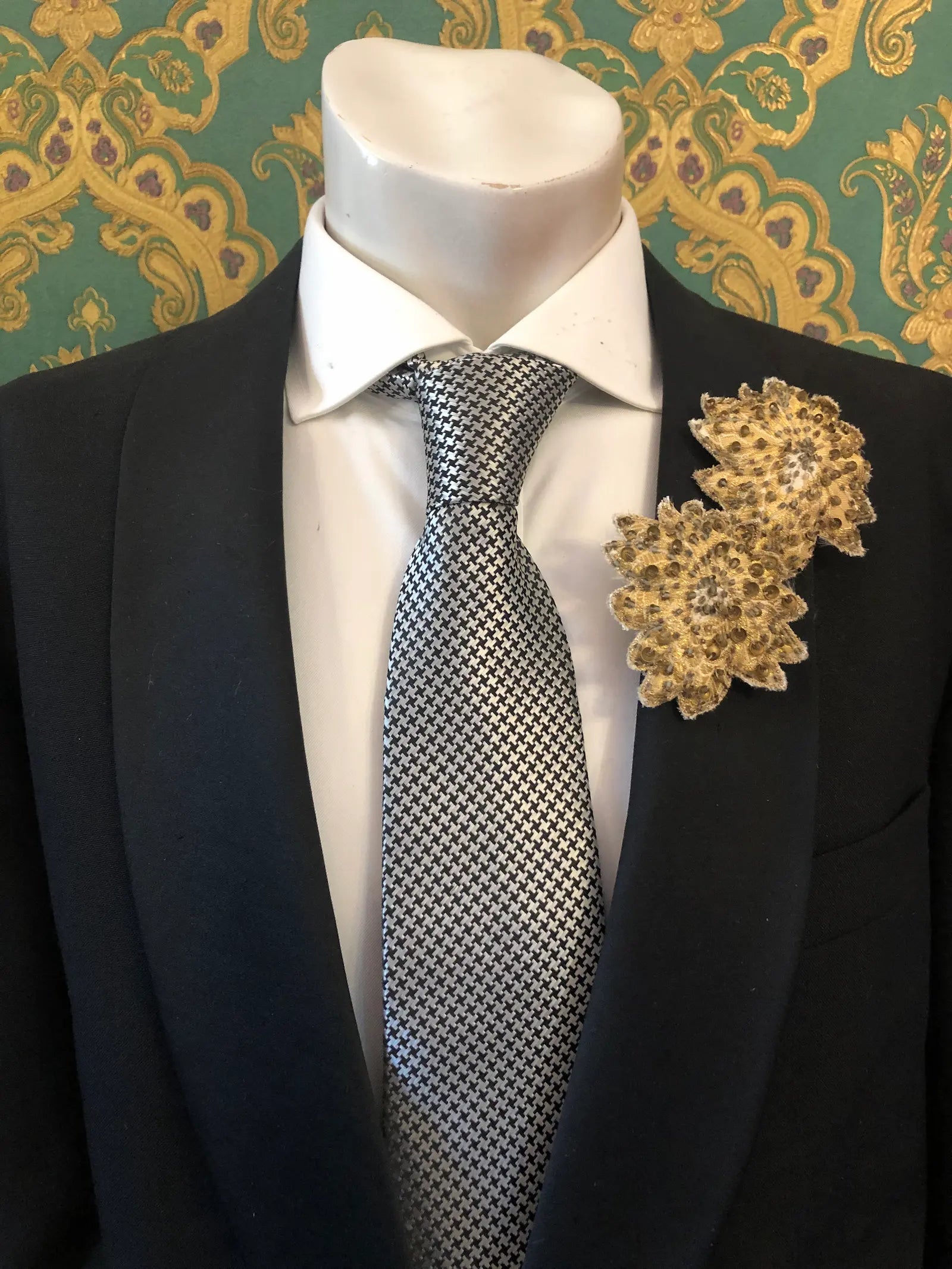 A mannequin torso wearing a black suit jacket, white dress shirt, patterned grey and white tie, and two Broochella Foglie d'autunno leaf brooches. The background is a green and damask-patterned wallpaper.