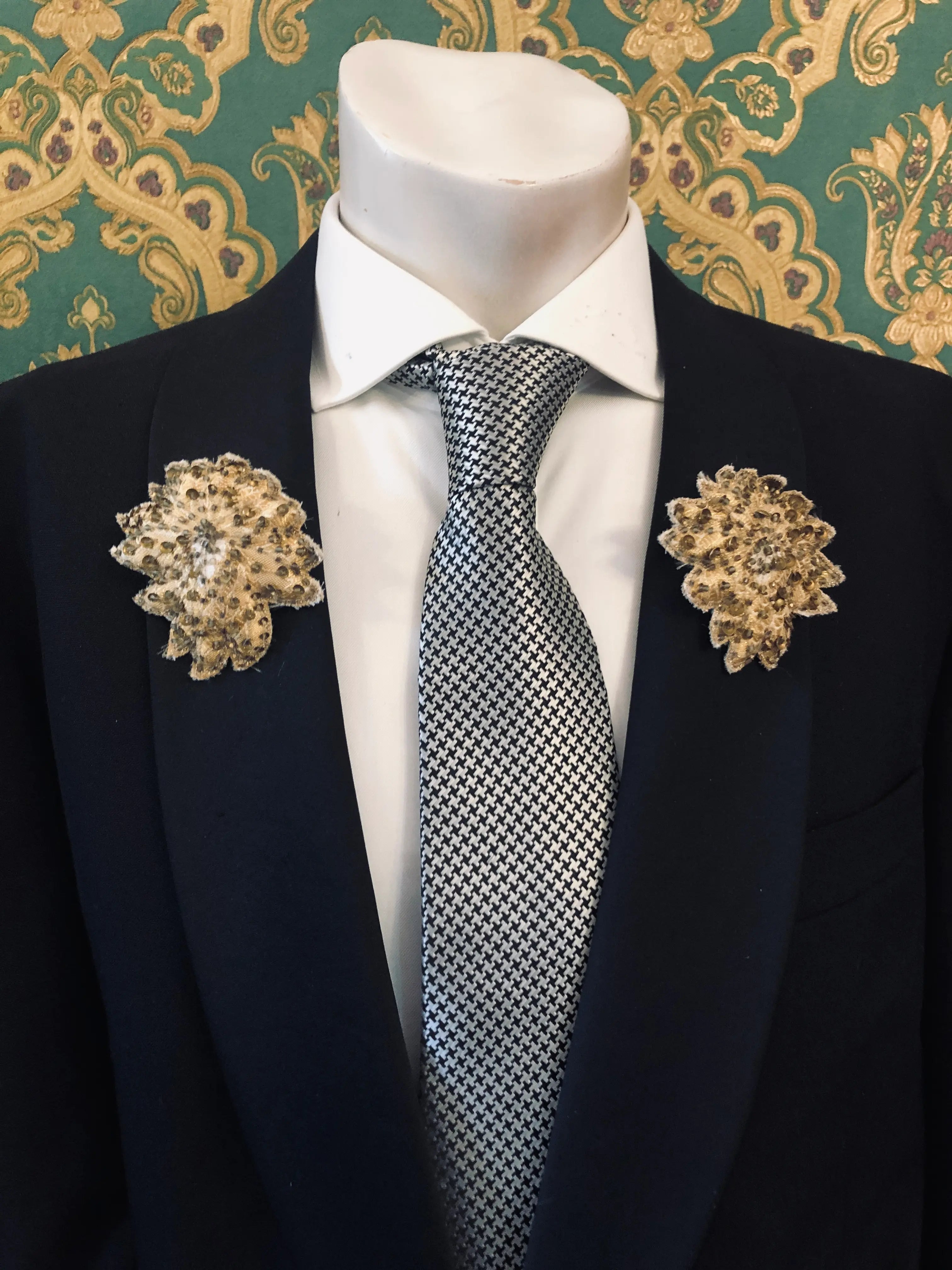 A mannequin torso wearing a black suit jacket, white dress shirt, patterned grey and white tie, and two Broochella Foglie d'autunno leaf brooches. The background is a green and damask-patterned wallpaper.