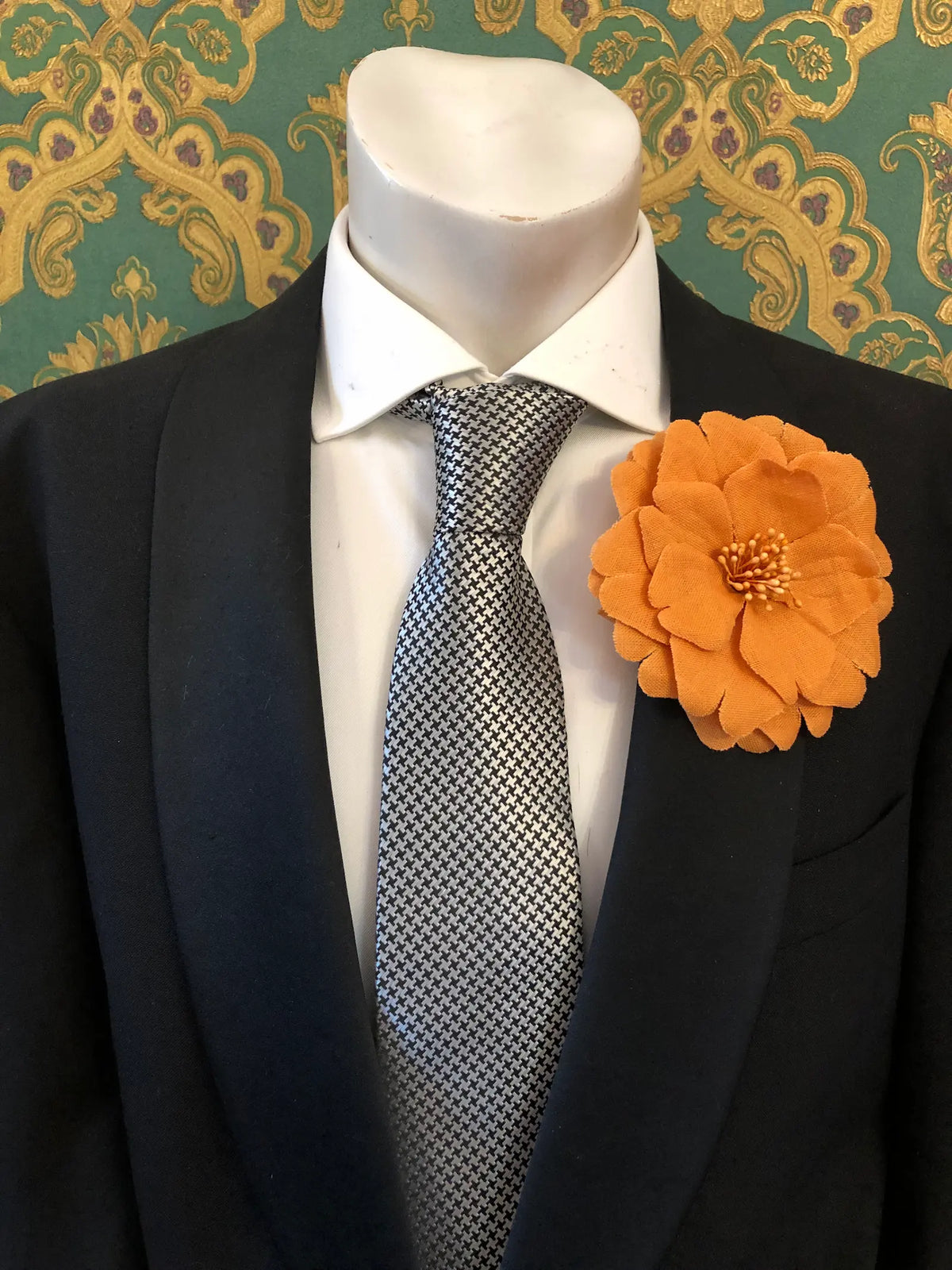 Broochella Men's Garofano handmade linen flower brooch in orange. It is pinned to a mannequin wearing a black suit, white shirt and a silver and black houndstooth tie. The background is green and gold damask wallpaper. 