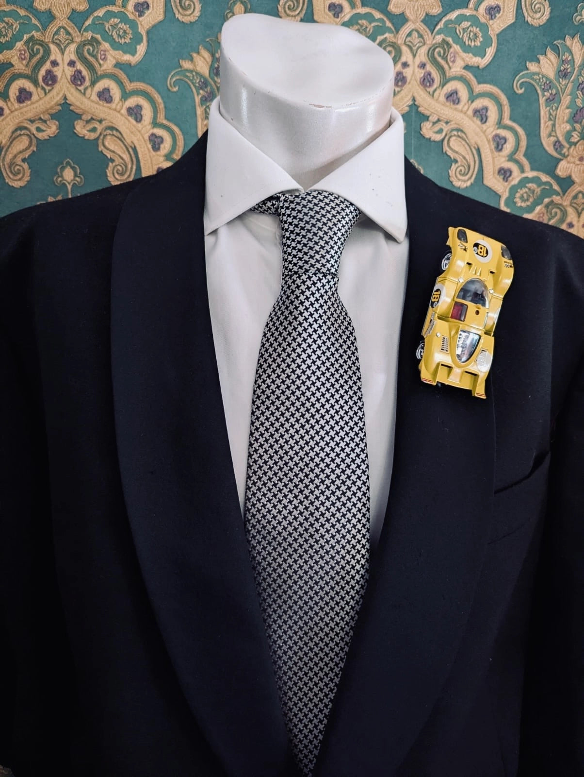 Broochella men's scuderia d'oro modena giallo yellow number 18 vintage formula one racing car brooch. Pinned on a men's mannequin wearing a black suit and a houndstooth tie. The background is green and gold damask wallpaper. 