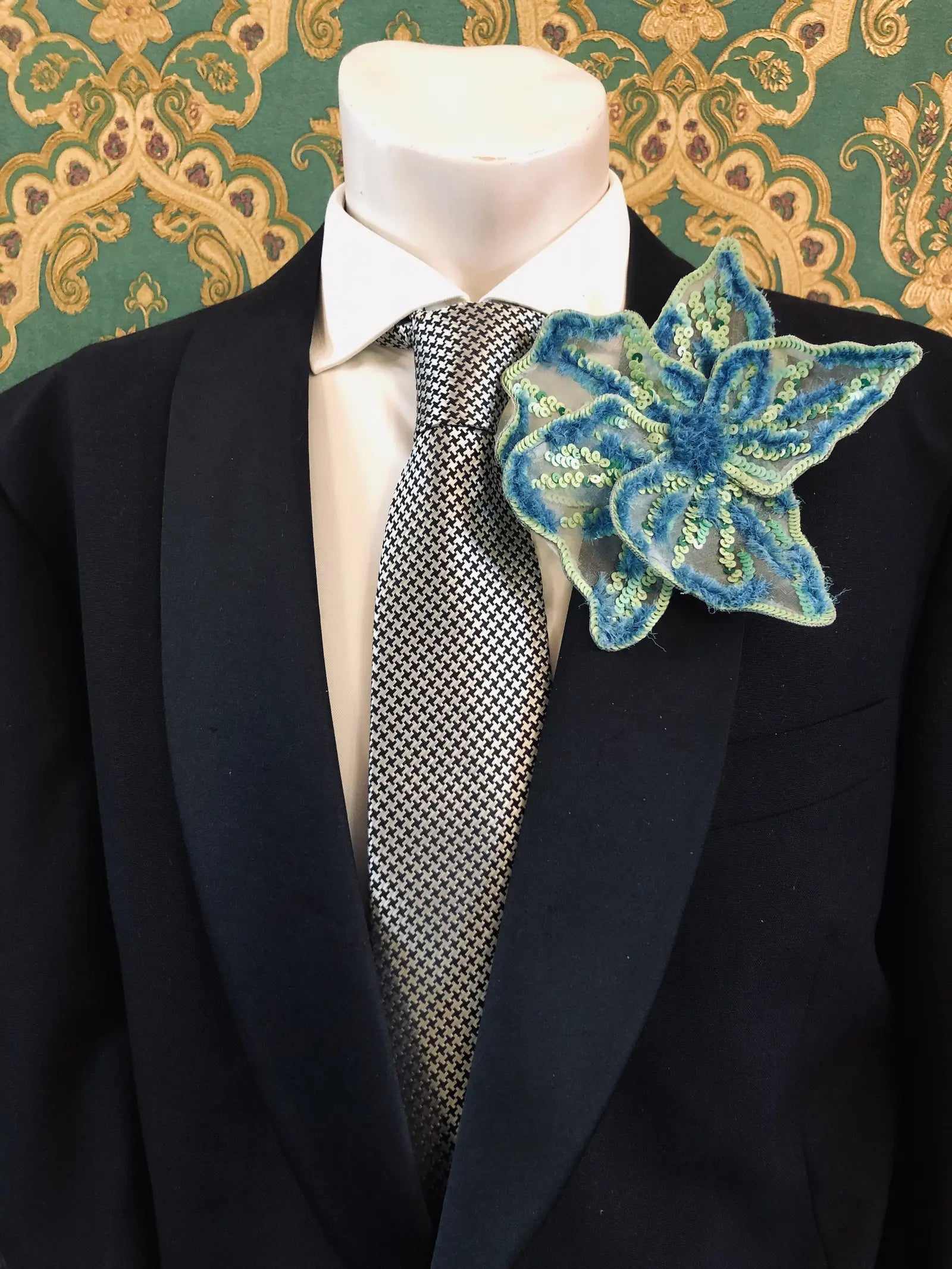A Broochella Men's Giglio di Tigre blue and green handmade sequinned  flower brooch pin. It is styled on a black suit jacket, white dress shirt, and patterned grey tie, set against a green and gold damask wallpaper background.
