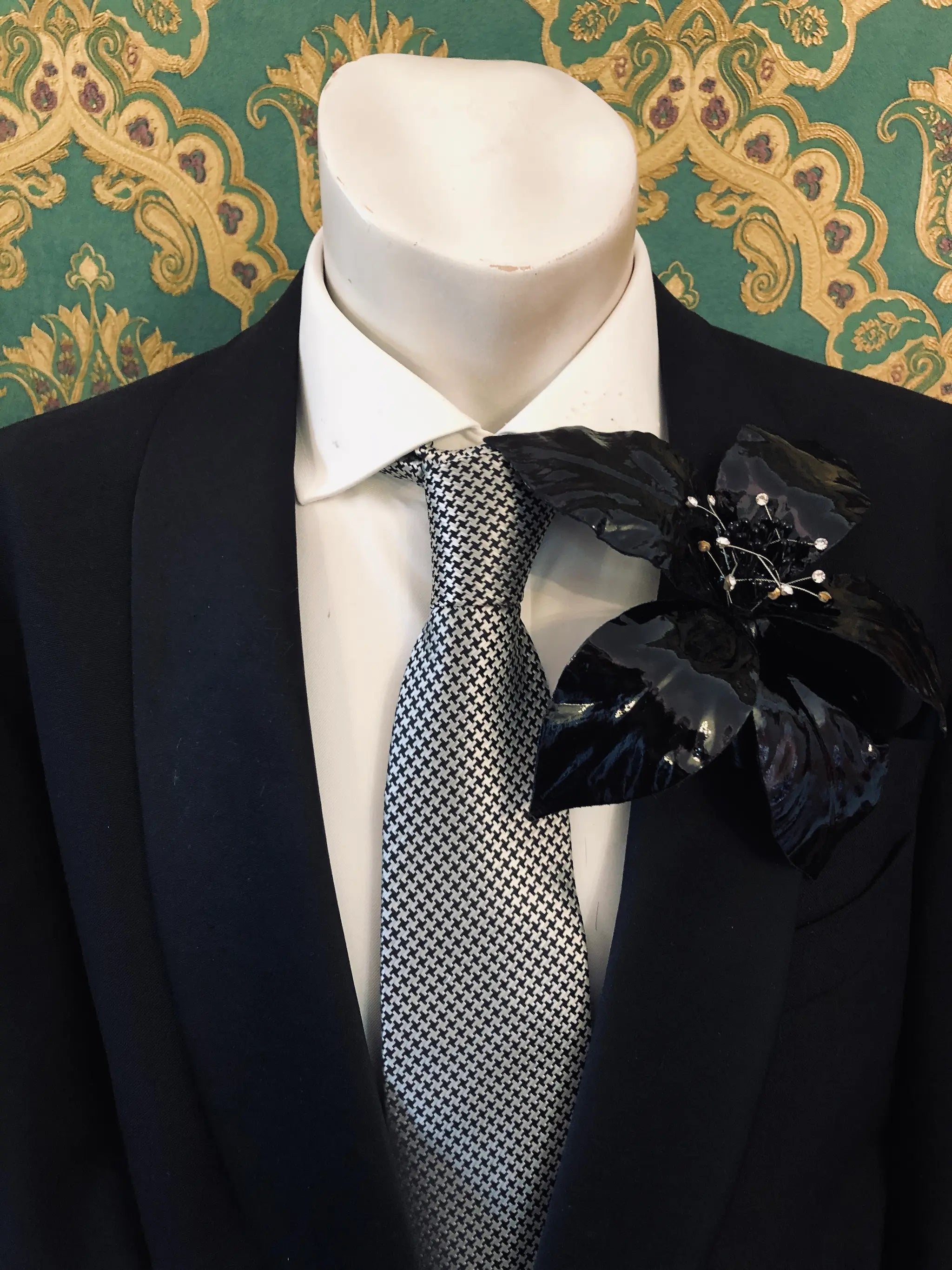Broochella Men's Giglio patent leather flower brooch in black. It is pinned to a men's mannequin that is dressed in a black suit with a grey and silver houndstooth tie. The background is green and gold damask wallpaper. 