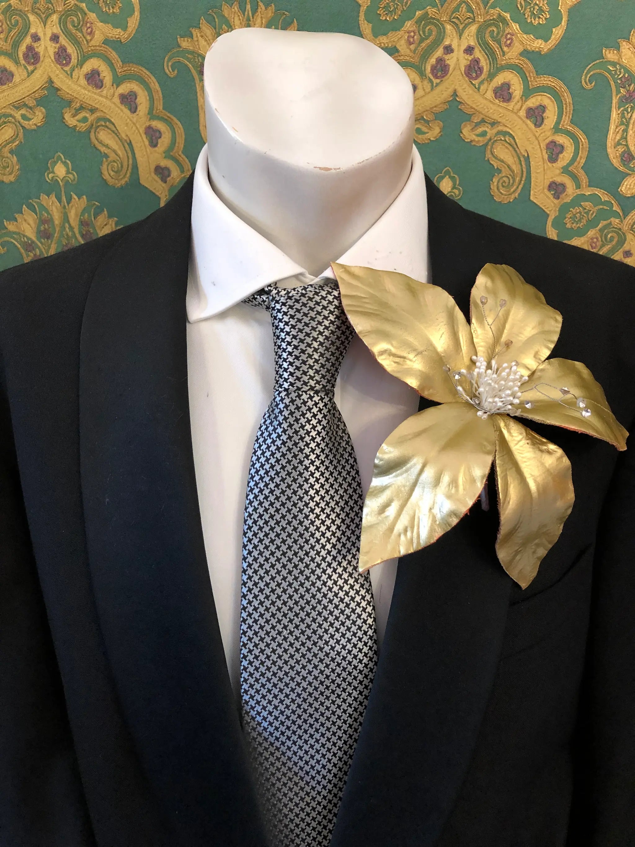 Broochella Men's Giglio patent leather flower brooch in gold. It is pinned to a men's mannequin that is dressed in a black suit with a grey and silver houndstooth tie. The background is green and gold damask wallpaper. 