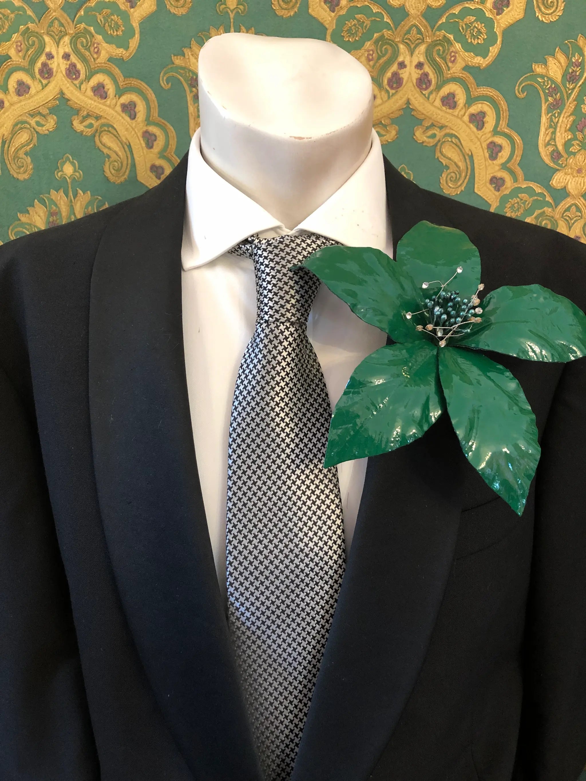 Broochella Men's Giglio patent leather flower brooch in green. It is pinned to a men's mannequin that is dressed in a black suit with a grey and silver houndstooth tie. The background is green and gold damask wallpaper. 