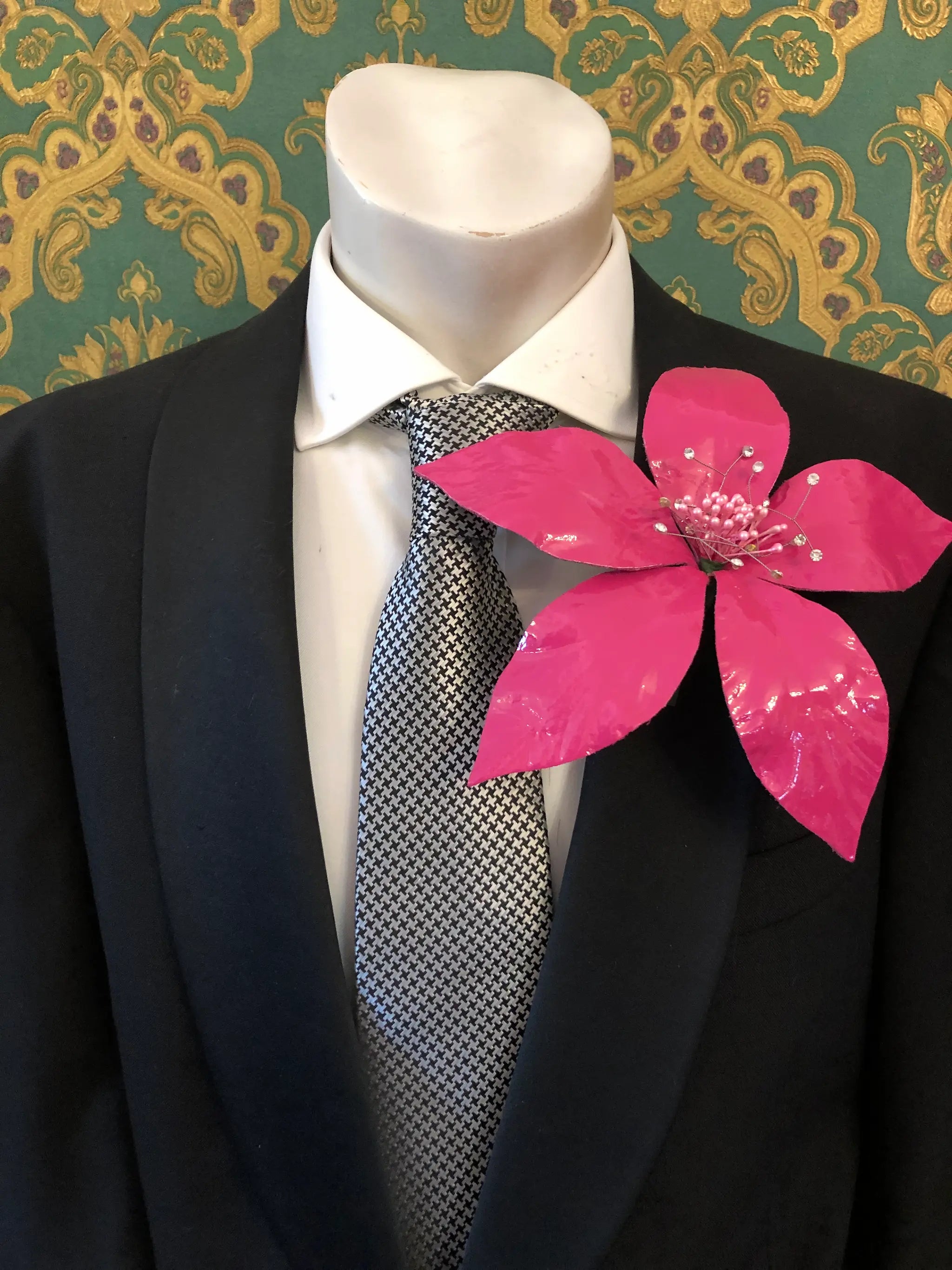 Broochella Men's Giglio patent leather flower brooch in pink. It is pinned to a men's mannequin that is dressed in a black suit with a grey and silver houndstooth tie. The background is green and gold damask wallpaper. 