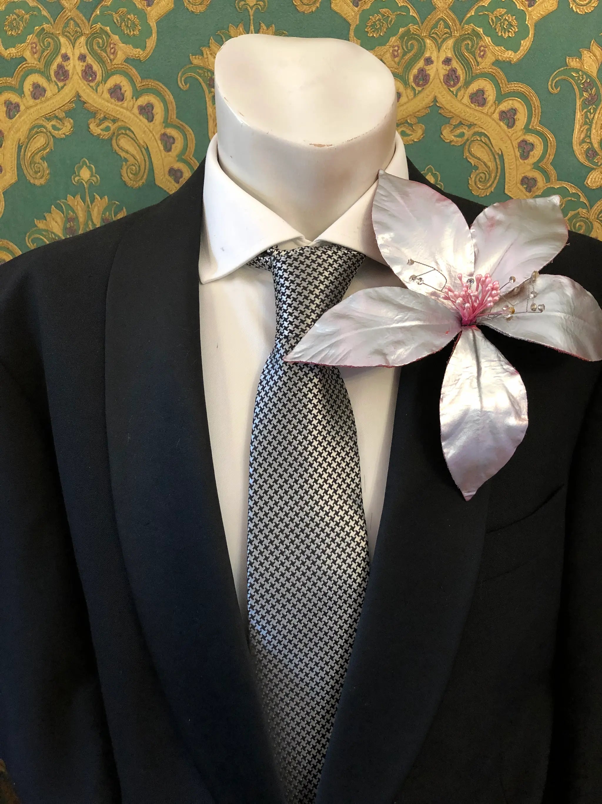 Broochella Men's Giglio patent leather flower brooch in silver. It is pinned to a men's mannequin that is dressed in a black suit with a grey and silver houndstooth tie. The background is green and gold damask wallpaper. 